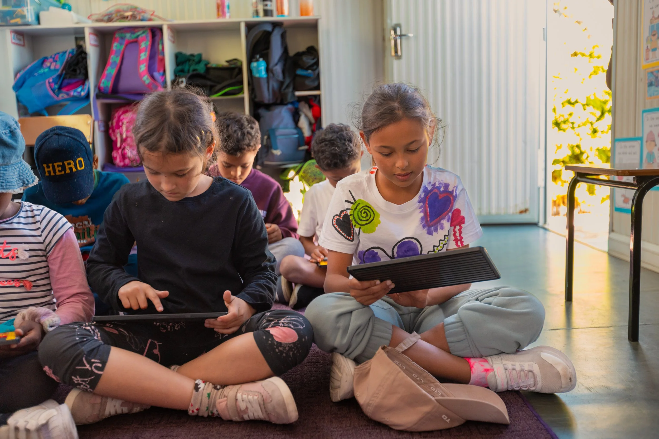 Group of children sitting on a carpeted floor, engaging with tablets indoors in a classroom setting with shelves and bright sunlight outside.