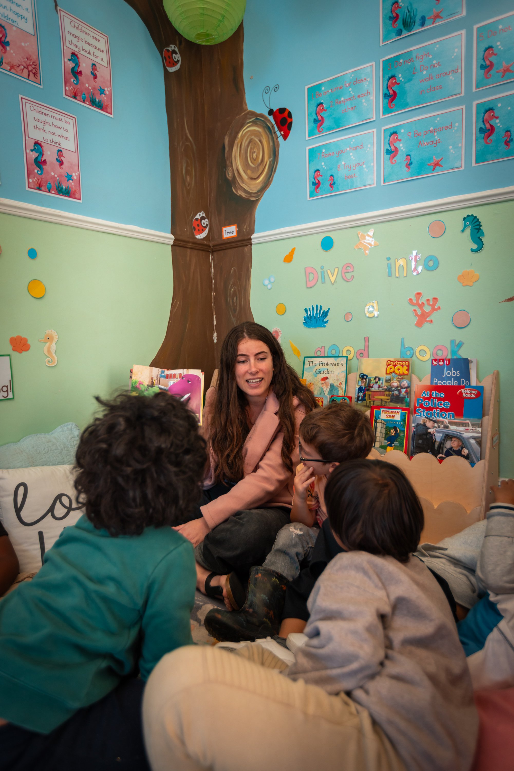 A classroom setting with a teacher and young students sitting on the floor. The background corner features a painted tree with seahorse and ladybug decorations, along with motivational rules written on blue posters. The wall below the posters has col