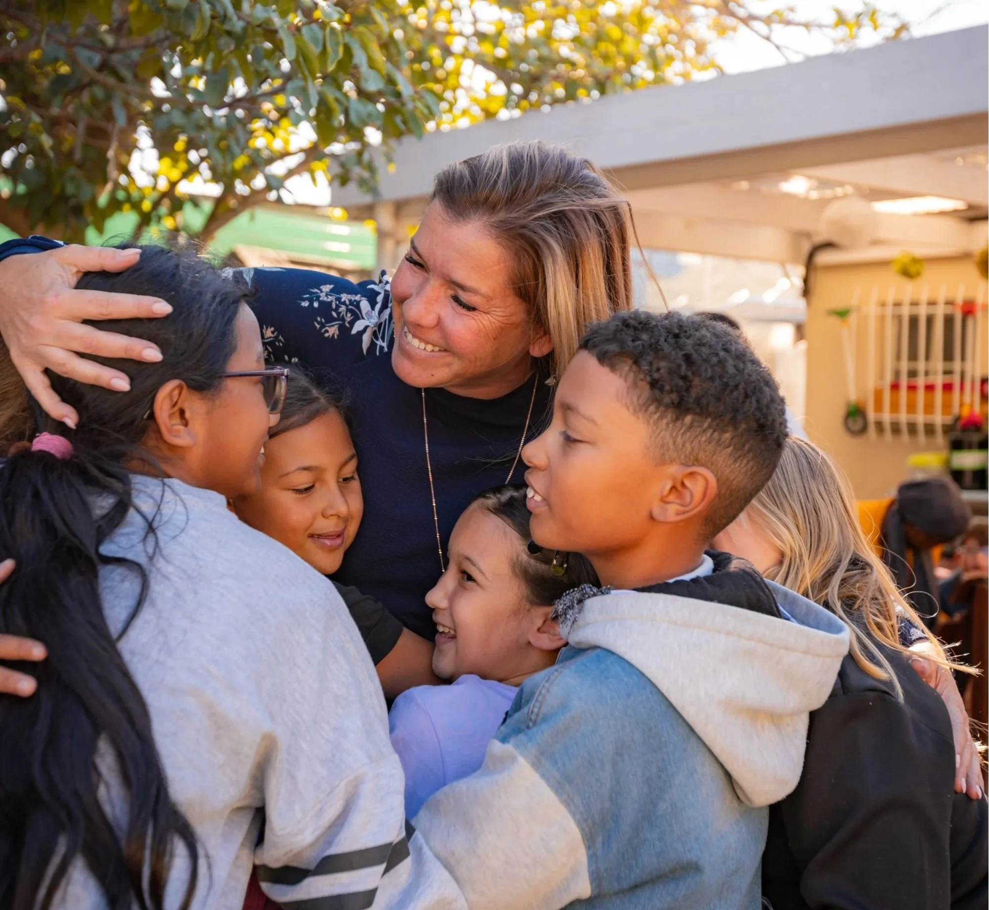 A group of children hugging a woman outdoors, possibly in a school or daycare setting, with trees and a structure in the background.