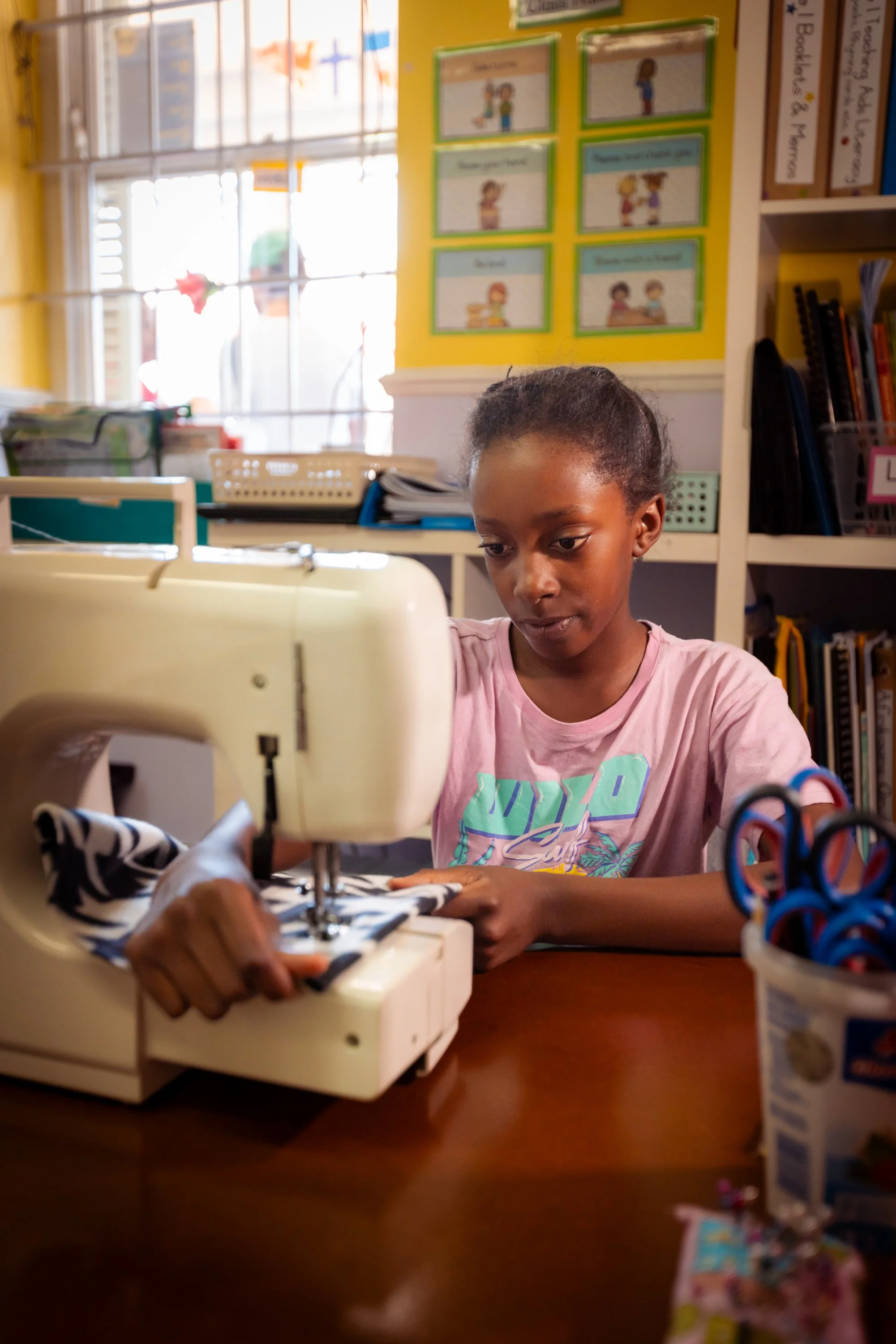 A young girl in a pink shirt working on a sewing project on a sewing machine in a classroom or craft room with bookshelves and posters in the background.