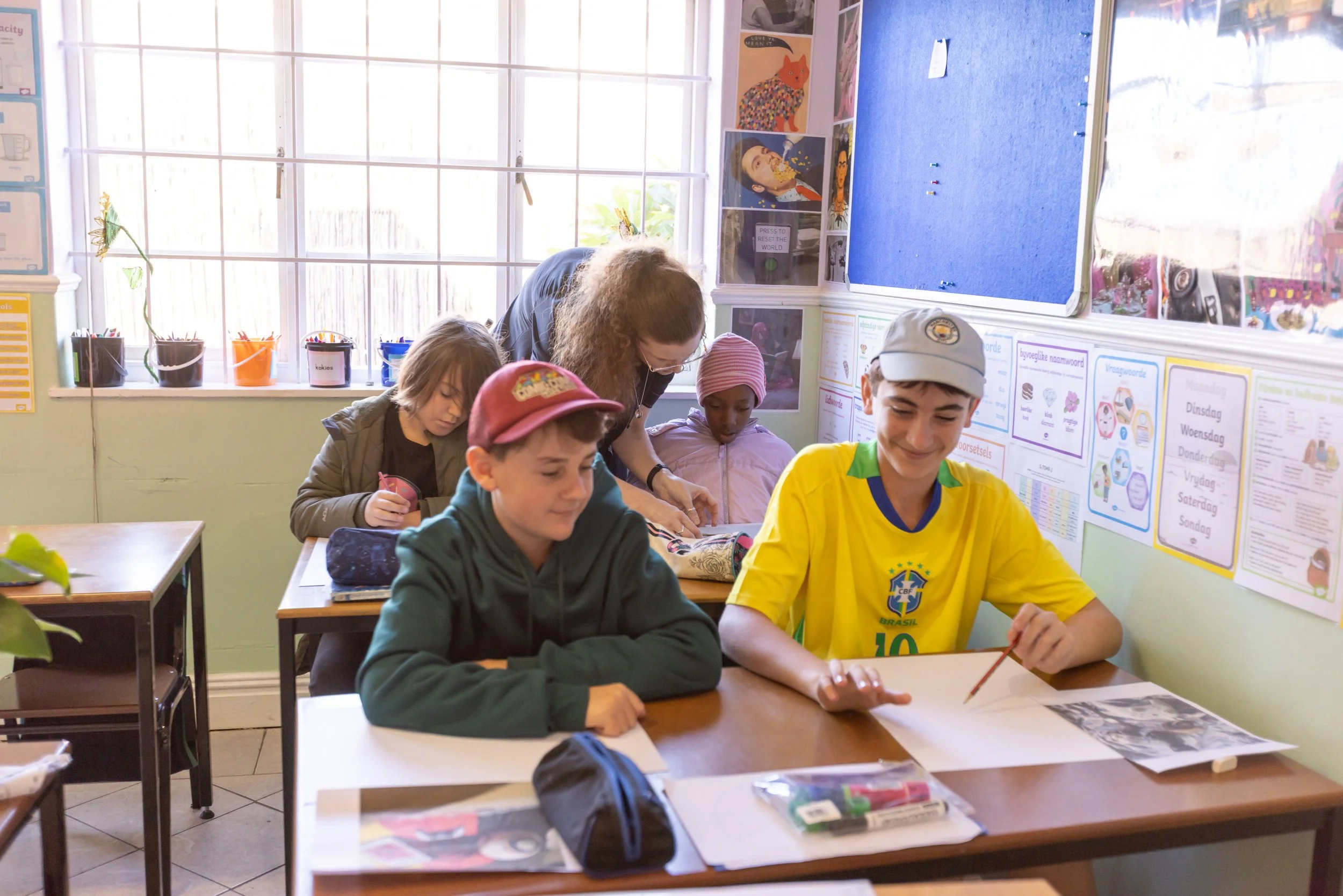 A classroom with students and a teacher seated at desks, working on assignments, with educational posters on the wall and a large window letting in natural light.