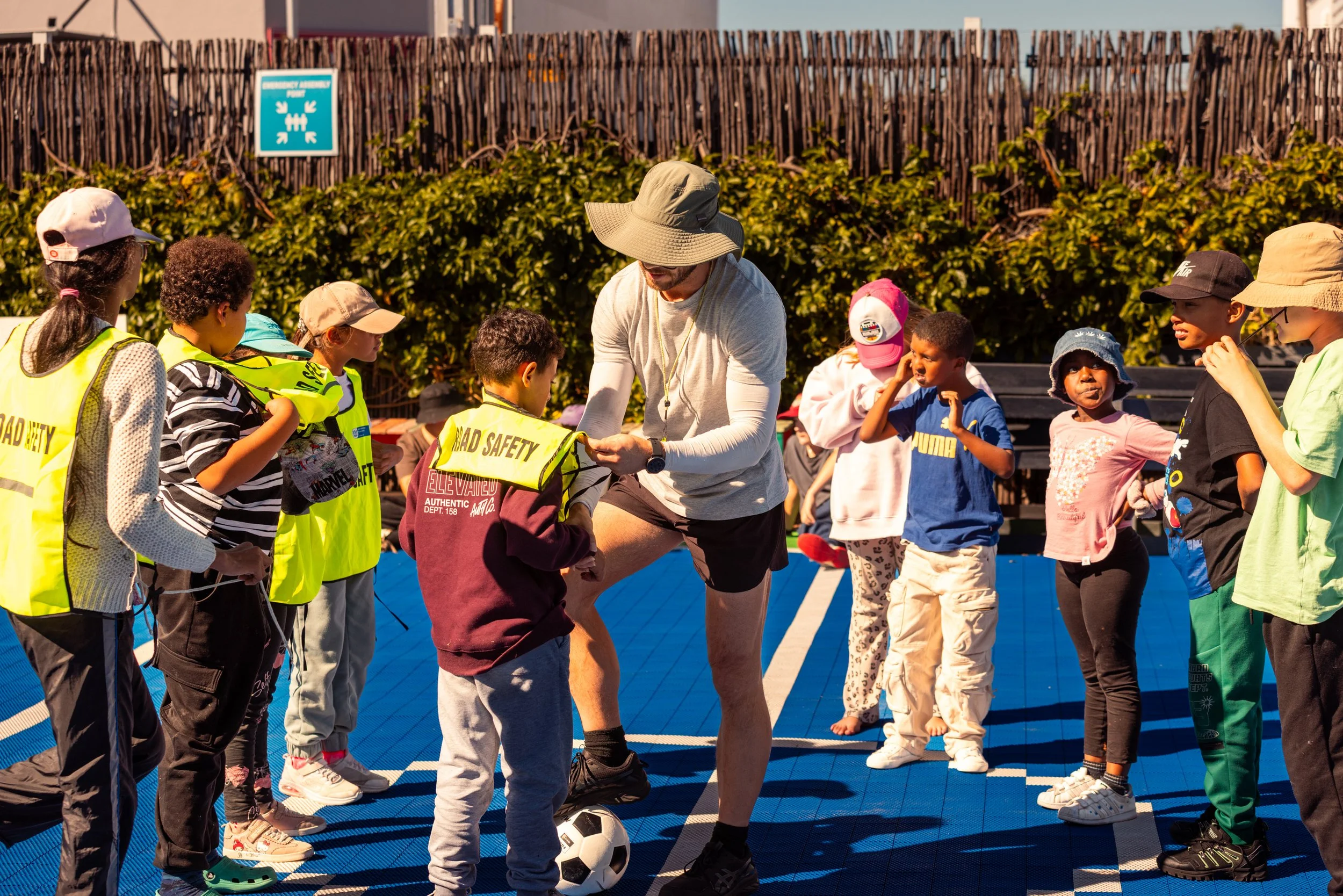 A group of children wearing yellow safety vests and hats, standing in a line on a blue surface, listening to an adult coach with a wide-brimmed hat and a beard, as they prepare for a soccer activity outdoors. The background shows a wooden fence and s