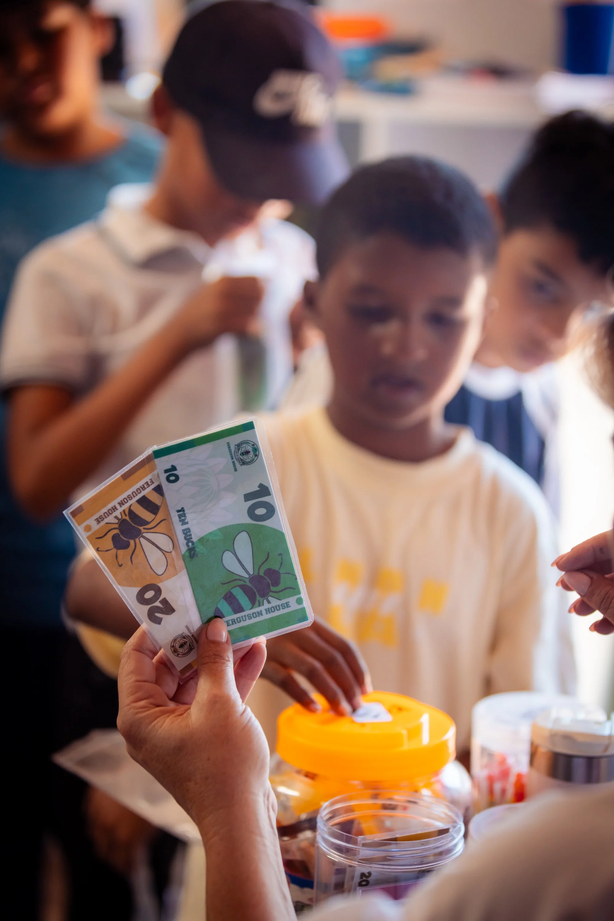 People participating in a voting or fundraising event, with one person holding two green $20 bills with a bee and flower design, and others putting money into collection containers on a table.