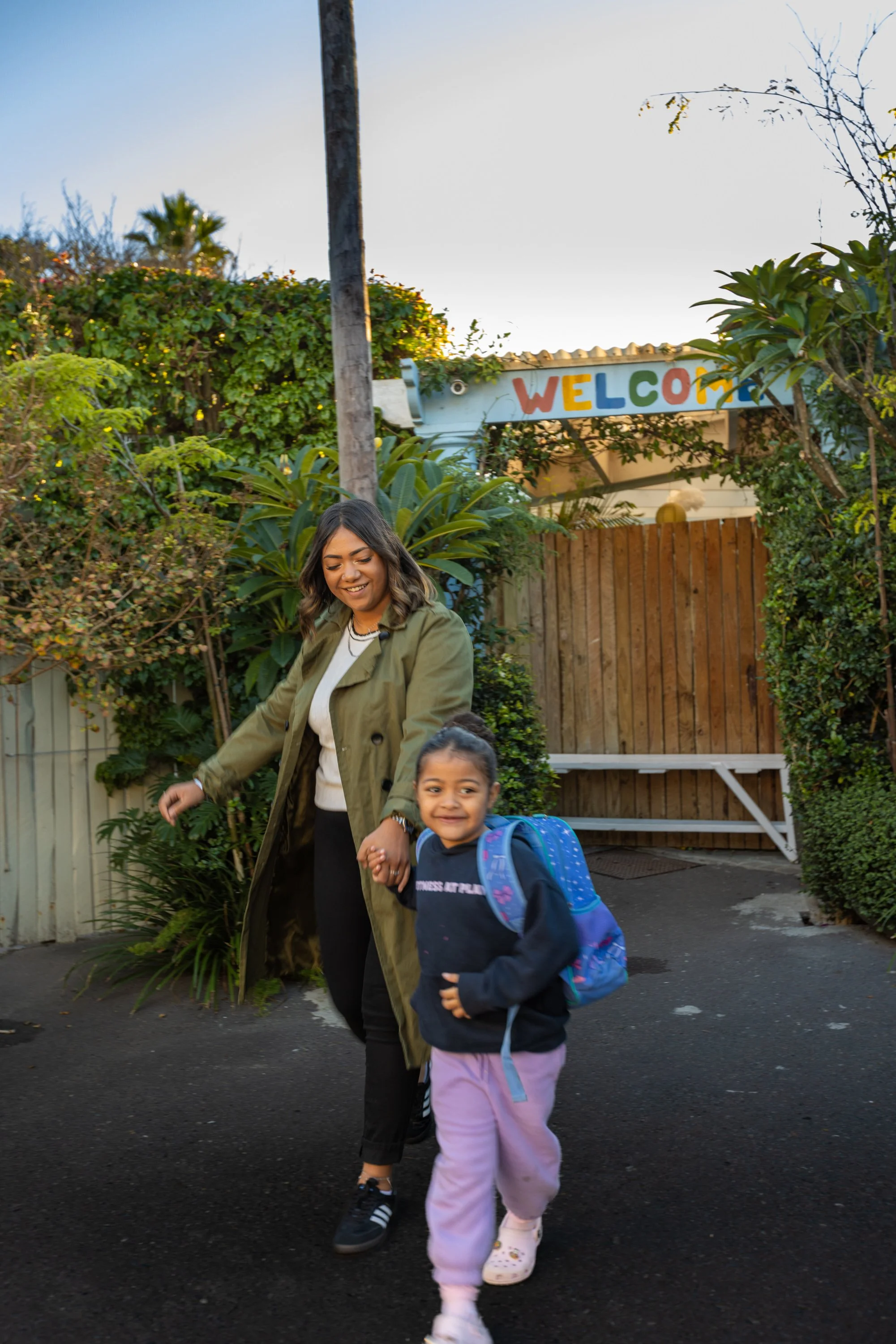 A woman and a young girl walking outdoors, holding hands, with the woman smiling and the girl looking happy. There is a colorful 