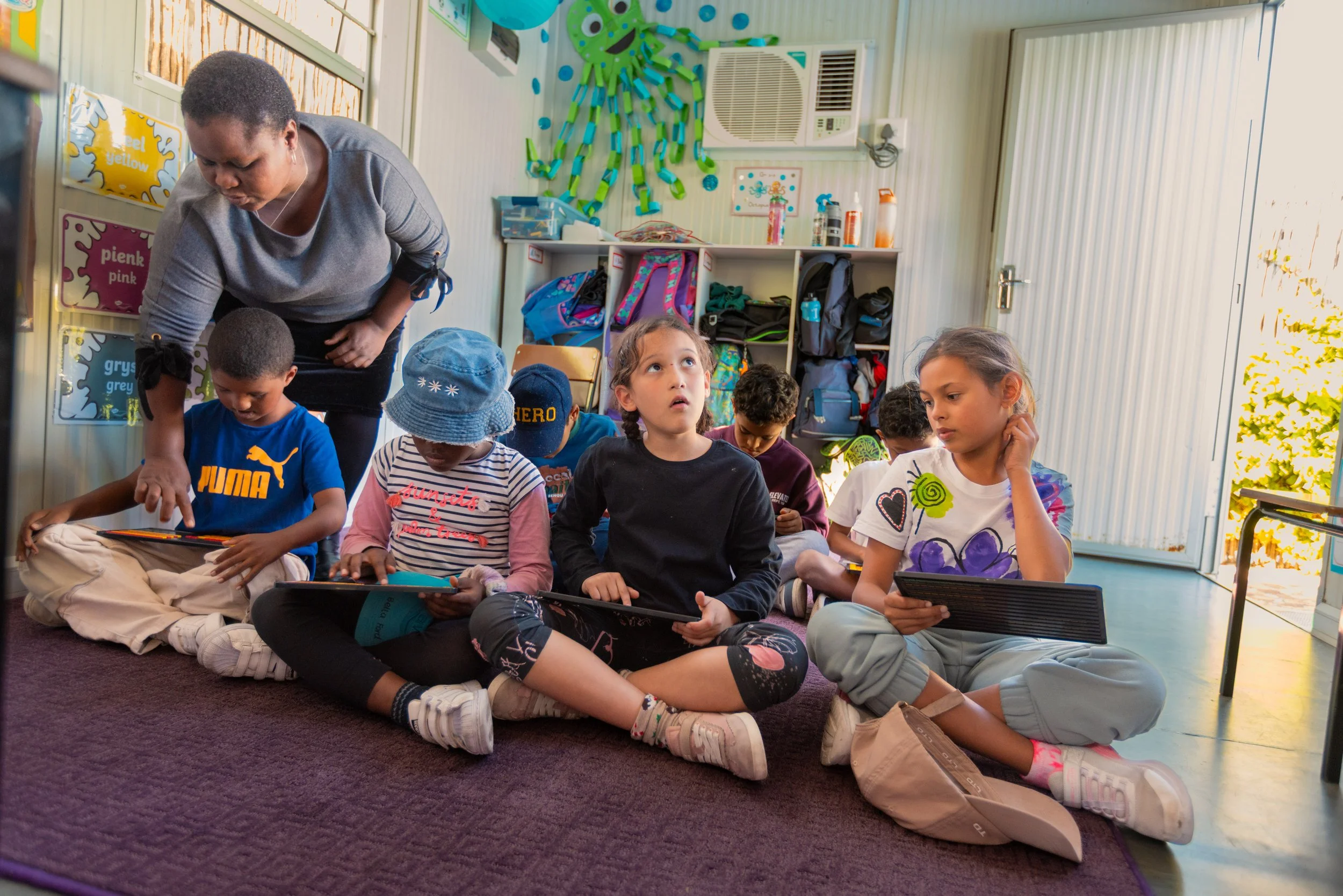 A group of young children sitting on the floor in a classroom, some playing on tablets or devices, with a teacher assisting. The classroom has colorful decorations and educational posters on the walls, and backpacks are stored in cubbies in the backg