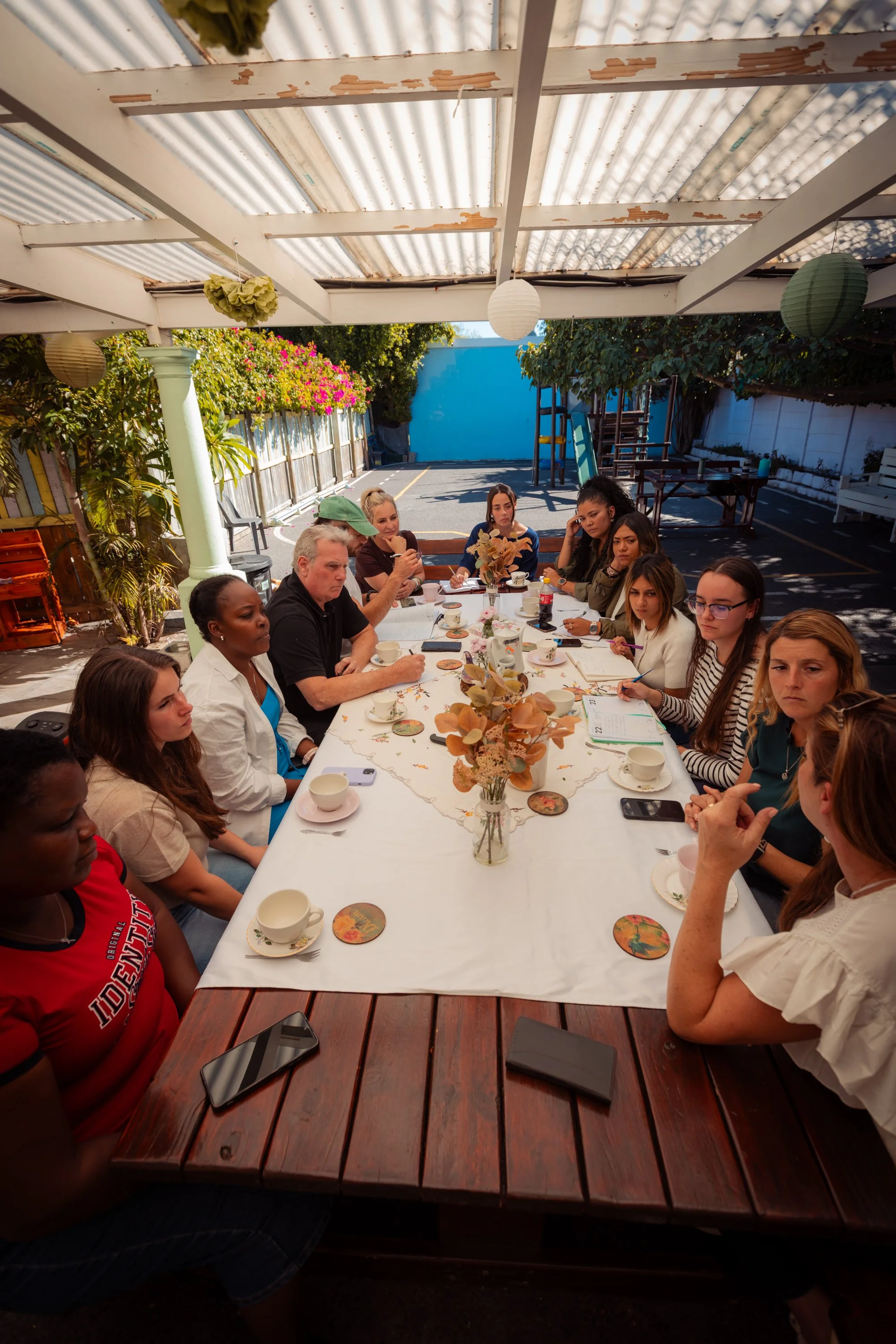 A group of people sitting around a long table outdoors, dining under a partially covered patio with hanging paper lanterns, foliage, and a blue wall in the background.