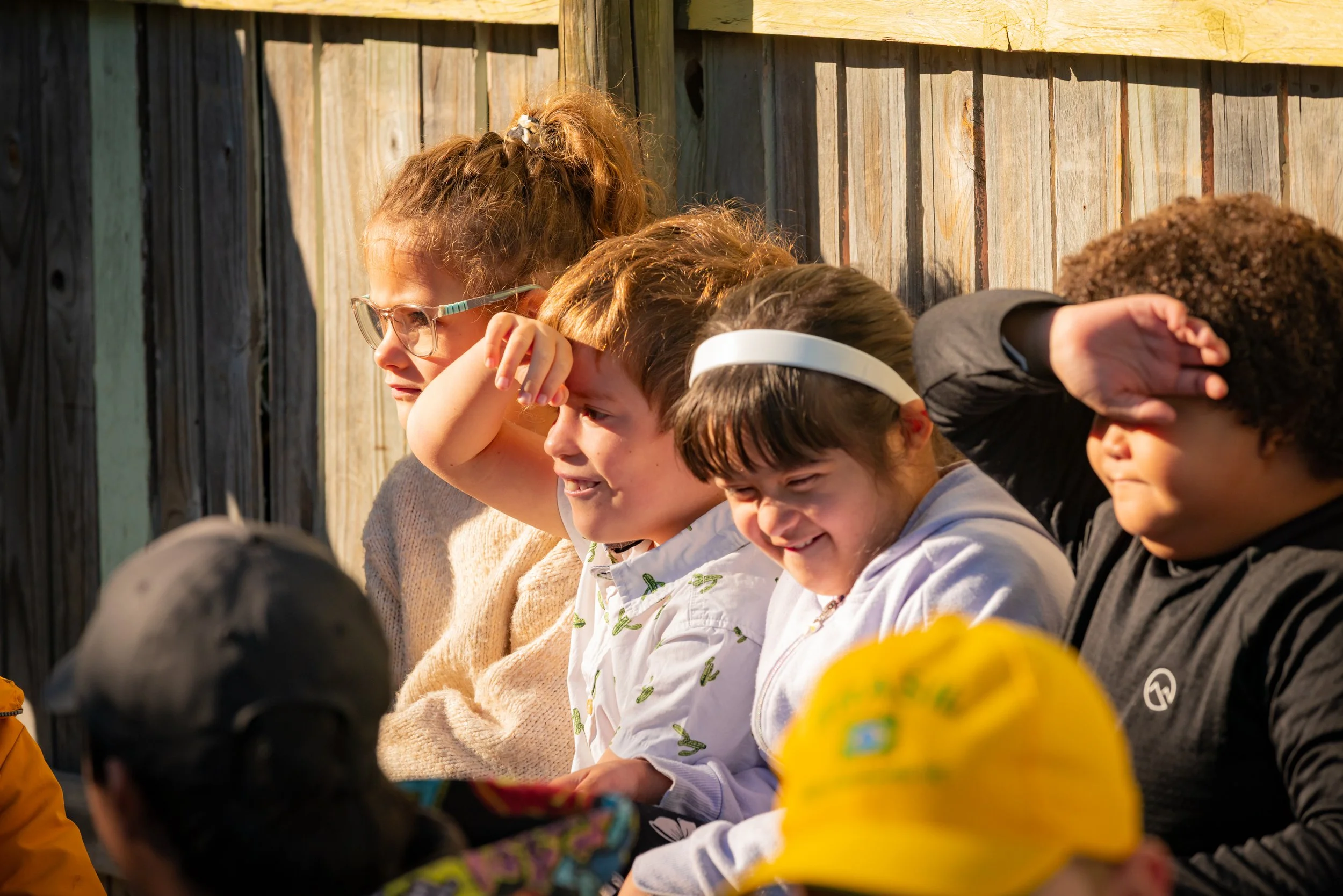 A group of children and a woman sitting outdoors, some shielding their eyes from the sun, next to a wooden fence.
