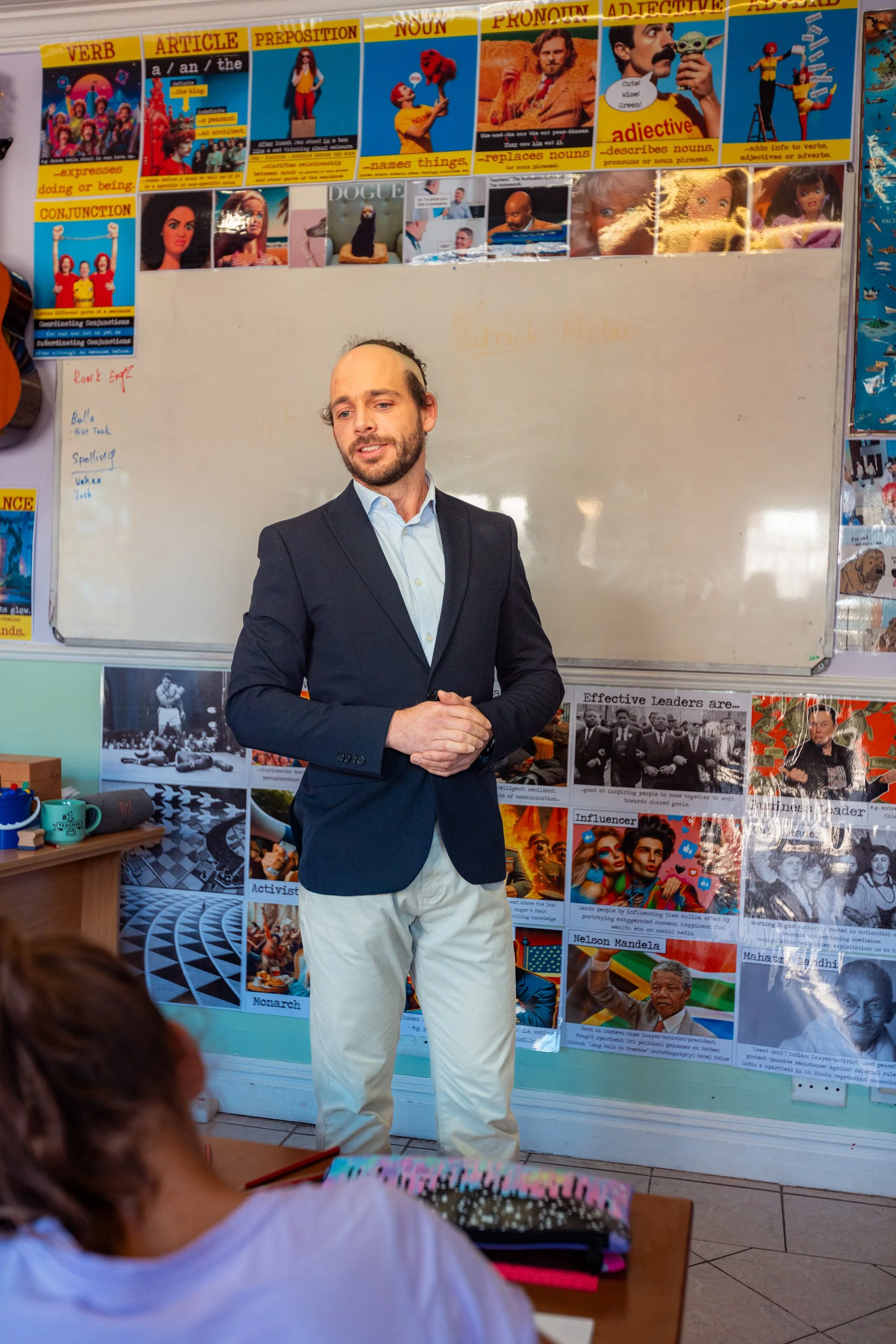 A man in a dark suit jacket and white pants standing in front of a whiteboard, giving a presentation in a classroom. The classroom walls are decorated with educational posters about grammar and influential figures.