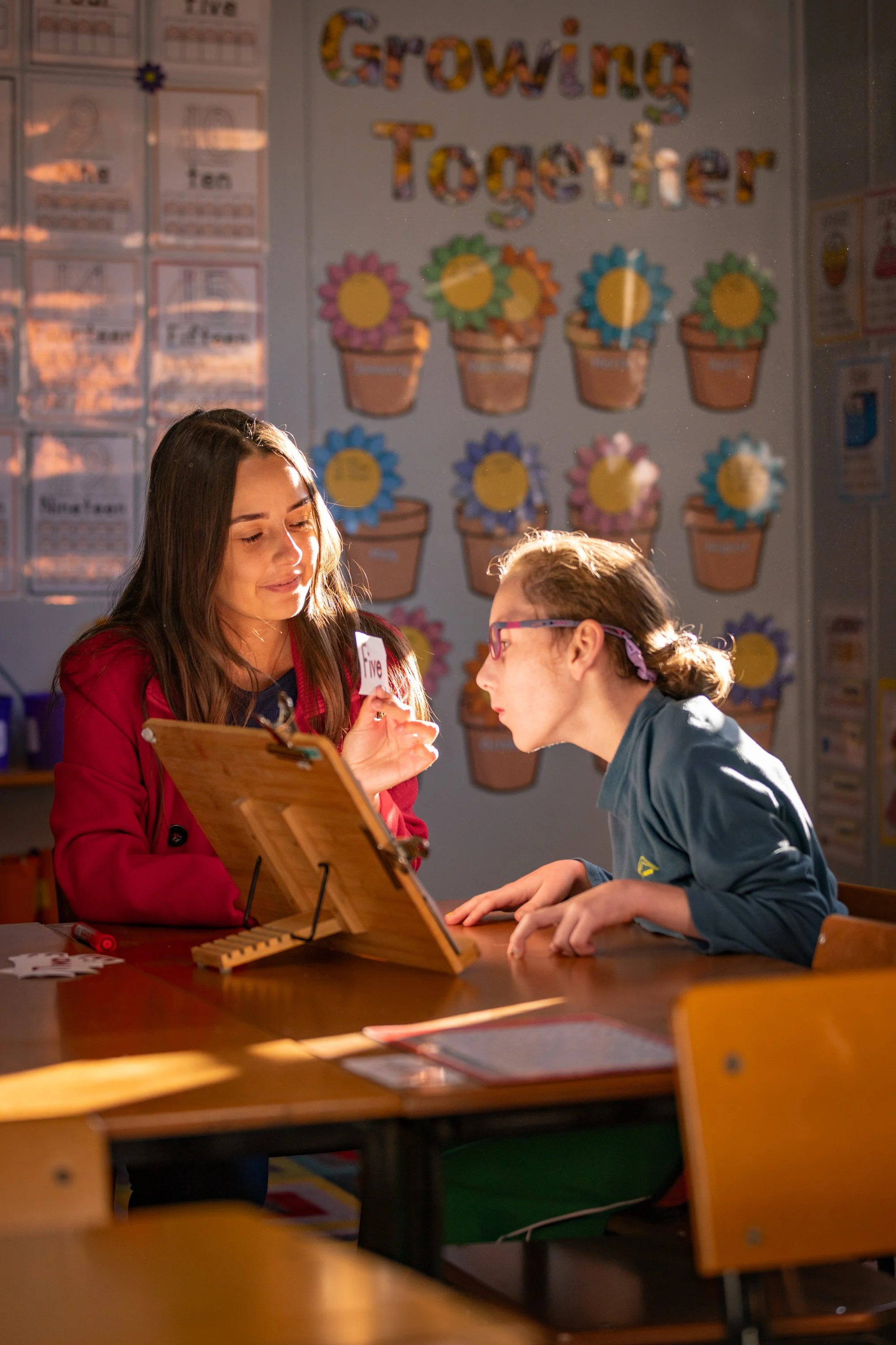 A teacher and a student are sitting at a desk in a classroom. The teacher is holding a card and smiling while the student looks engaged. Behind them, colorful flower decorations and a banner that says "Growing Together" are on the wall. There are educational posters and materials around the classroom.