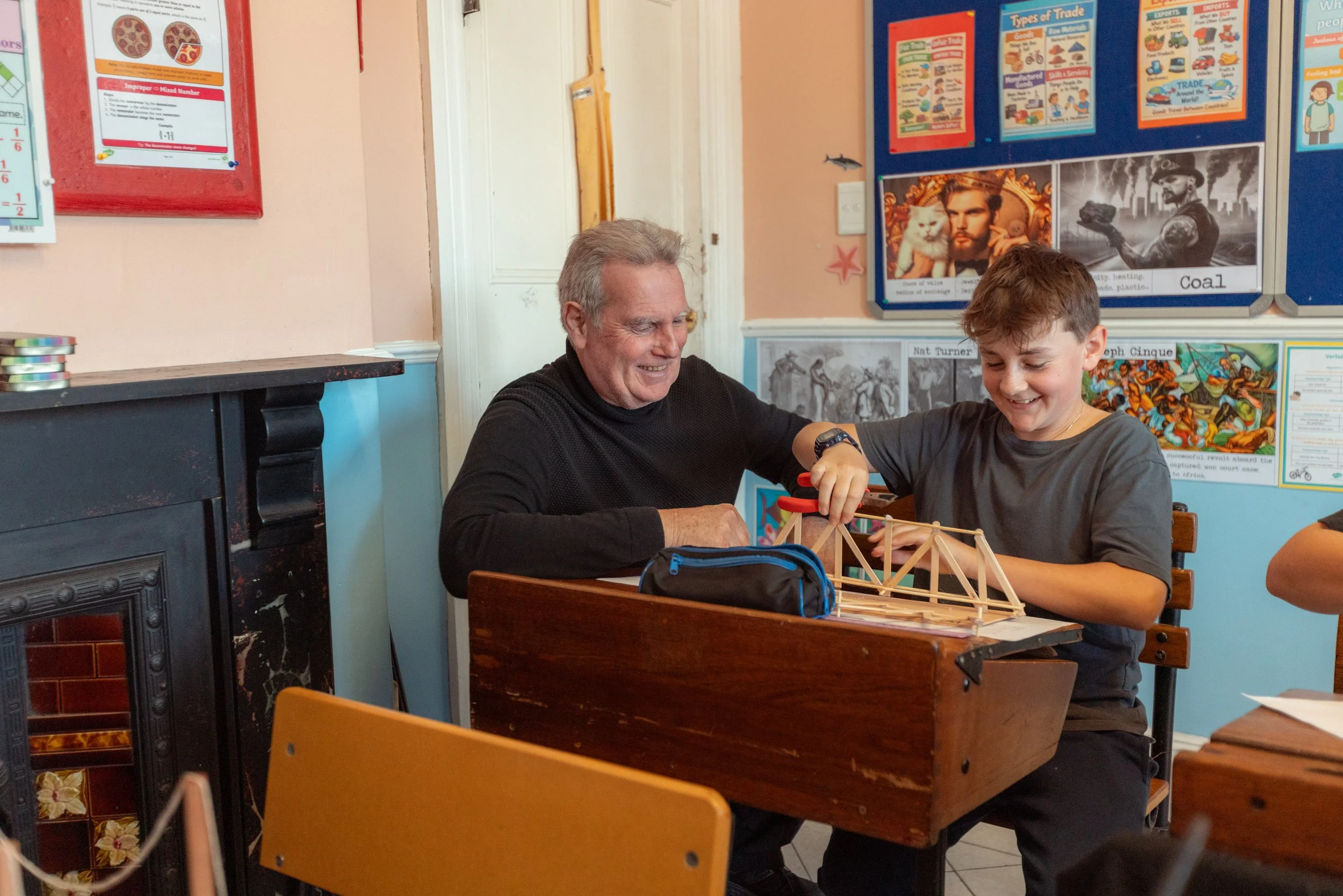 An elderly man and a young boy are playing with a wooden bridge model at a table in a classroom with educational posters and artwork on the wall.