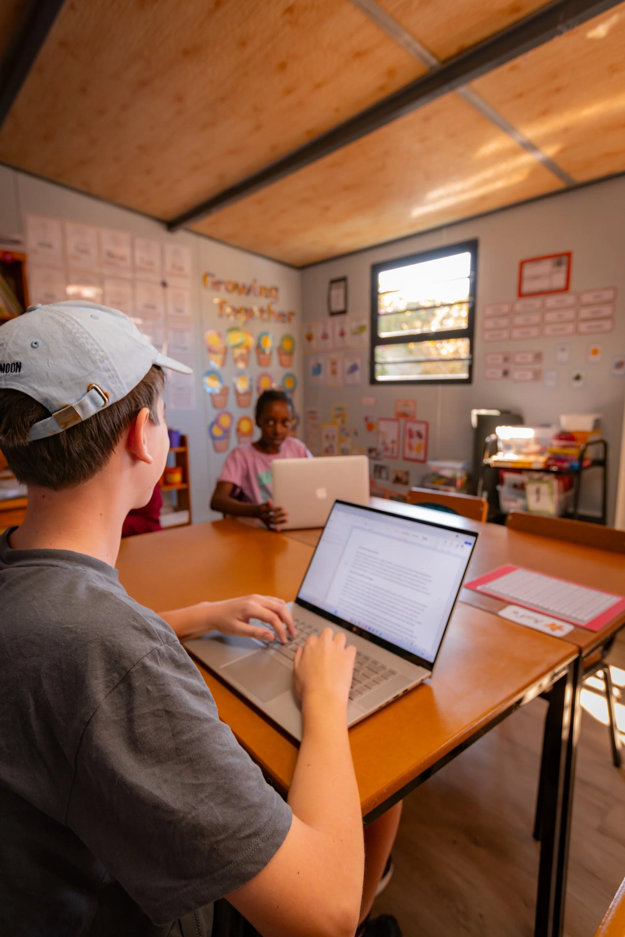 A young boy with a white cap typing on a laptop in a classroom with a girl in the background, educational posters on the walls, and a window letting in natural light.