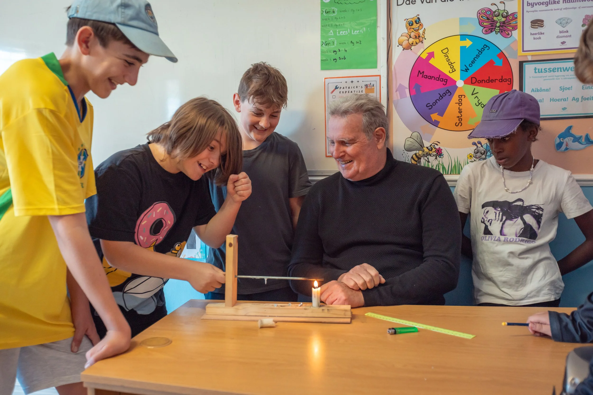 A group of children and an adult gather around a table with a lit candle and a small wooden device. They are engaging in a science activity, observing the flame. The background features educational posters with cartoon characters and schedule notes on a classroom wall.