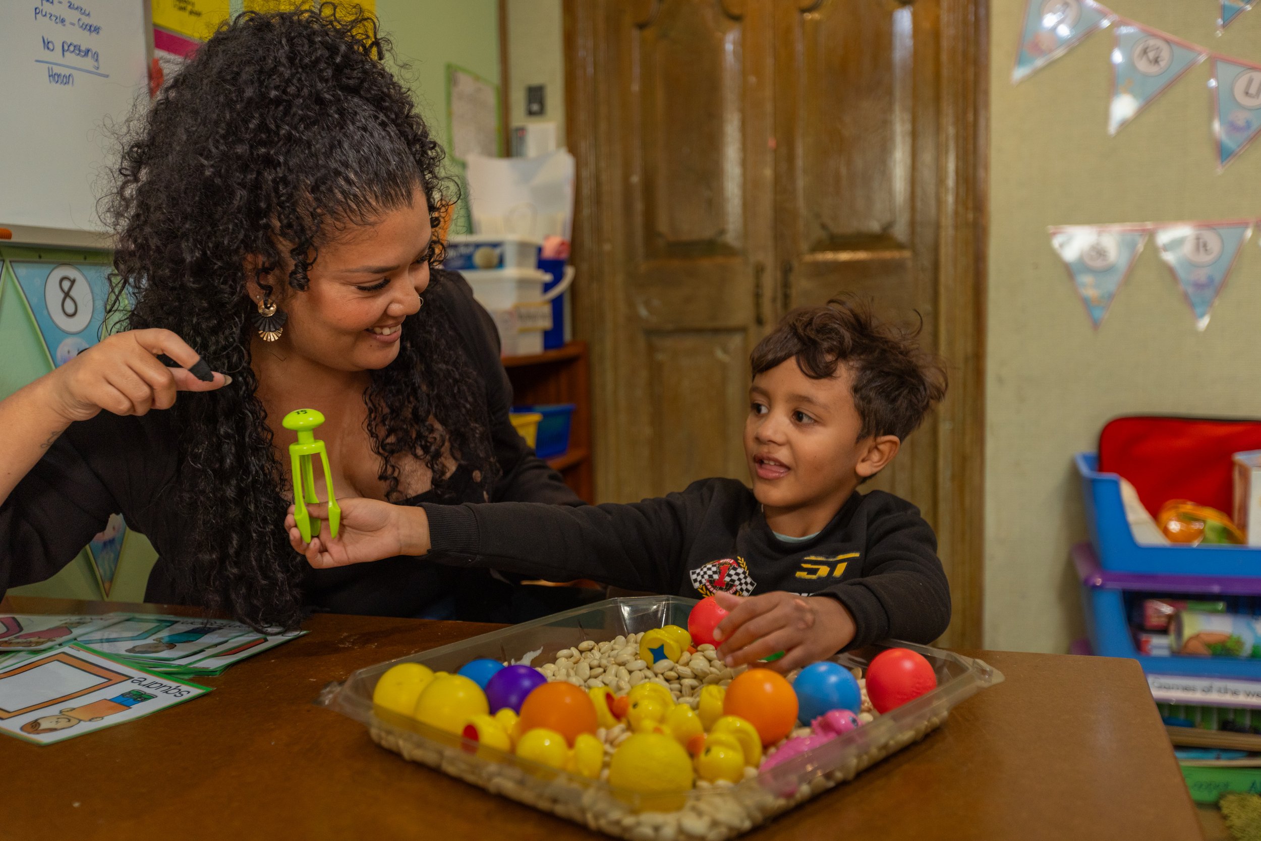 A woman and a young boy are sitting at a table playing with colorful plastic balls. The woman is smiling and holding a green ball, the boy is reaching for a red ball. The table has a tray of assorted plastic balls and some game cards. The background 