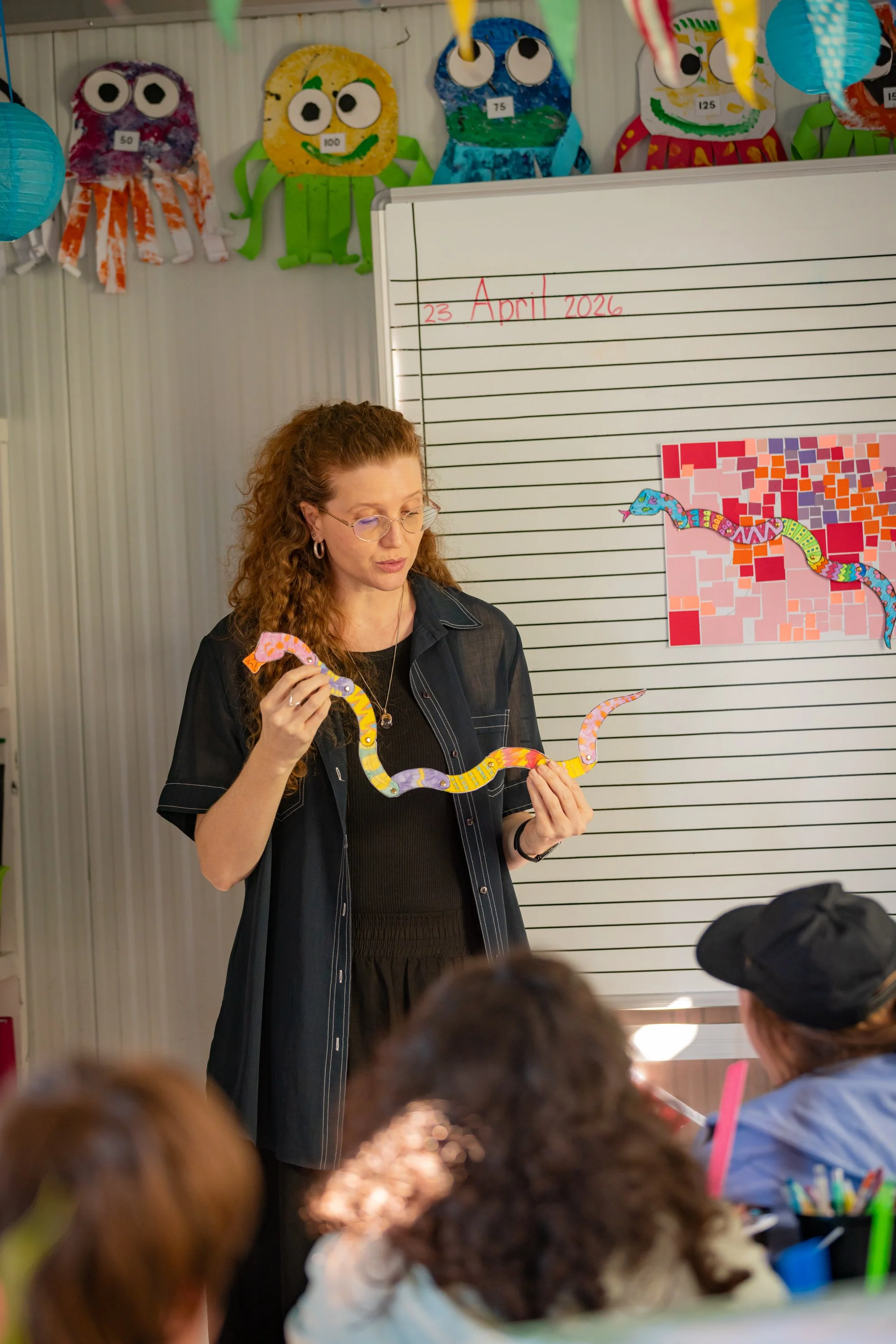 A woman with curly red hair, glasses, and earrings standing in front of a whiteboard with red and black writing that shows the date "23 April 2026." She is holding a colorful paper snake craft. Several children are seated, some visible in the foregro