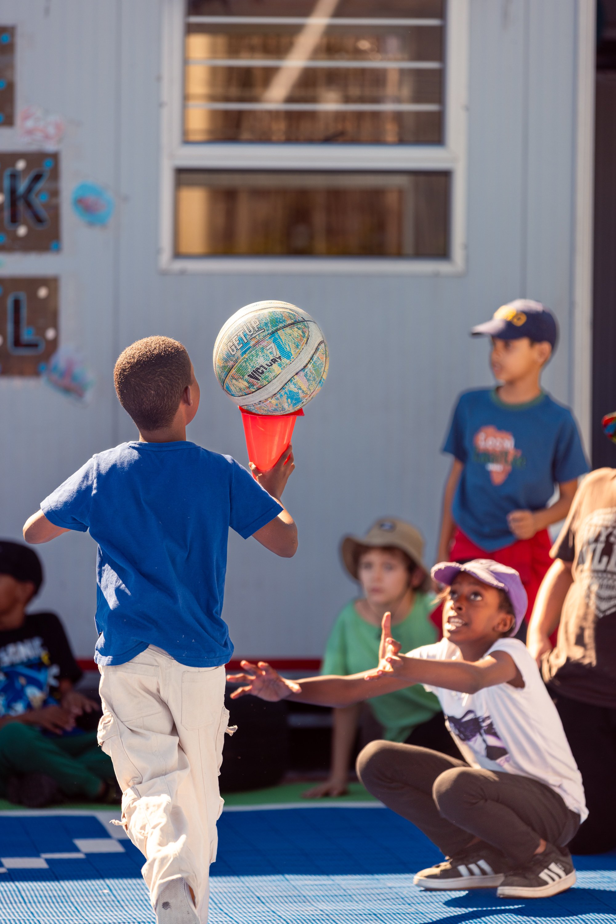 A young boy in a blue shirt and white pants is playing basketball outdoors while a group of children and adults watch and cheer.