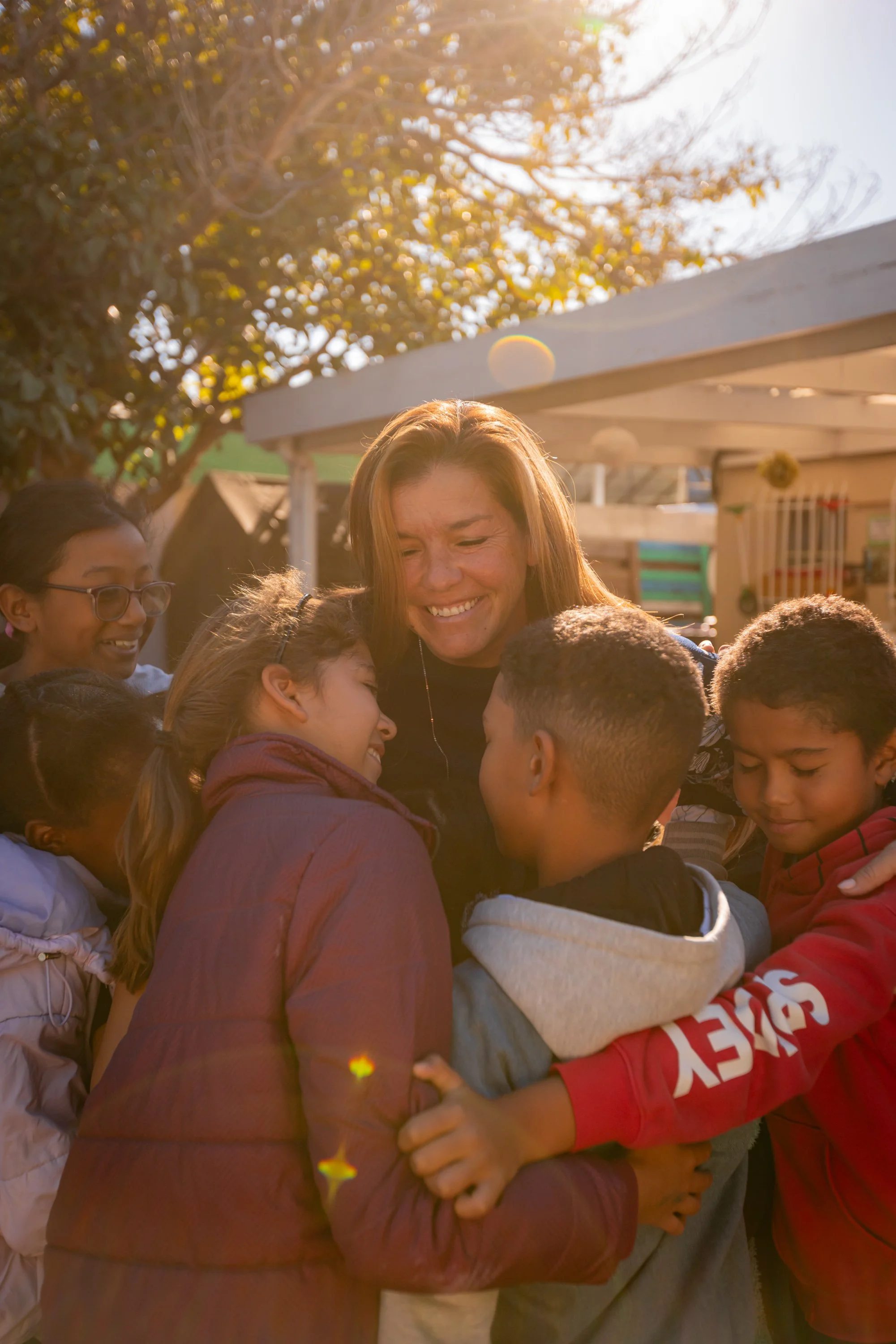 A group of people, including children and adults, are huddled together outdoors in a circle, smiling and embracing each other in a warm, sunny setting with trees in the background.