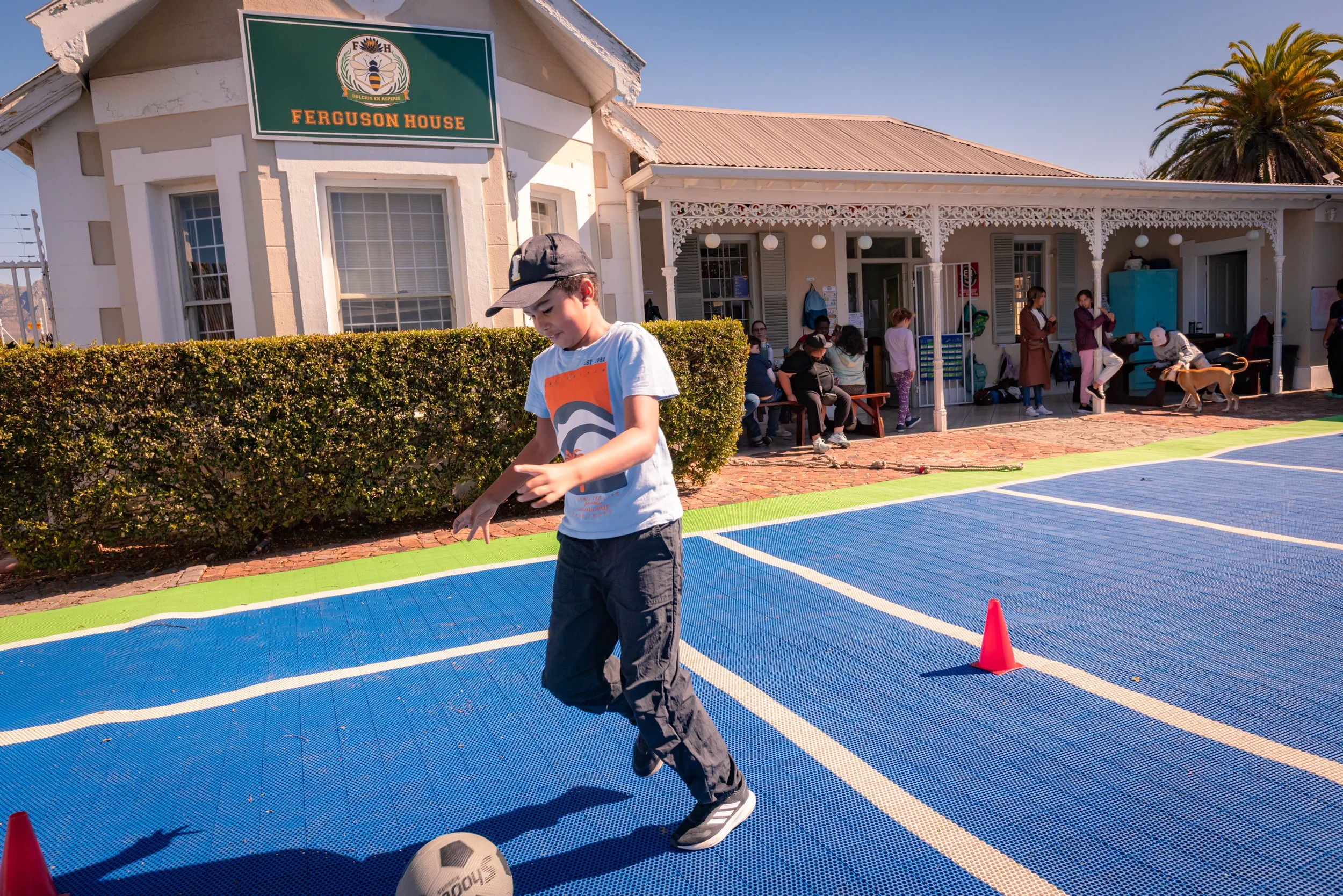 A young boy in a white t-shirt and black pants playing soccer on a blue and green court with orange cones, outside Ferguson House which features a white Victorian-style building with a porch filled with people and a large palm tree in the background.