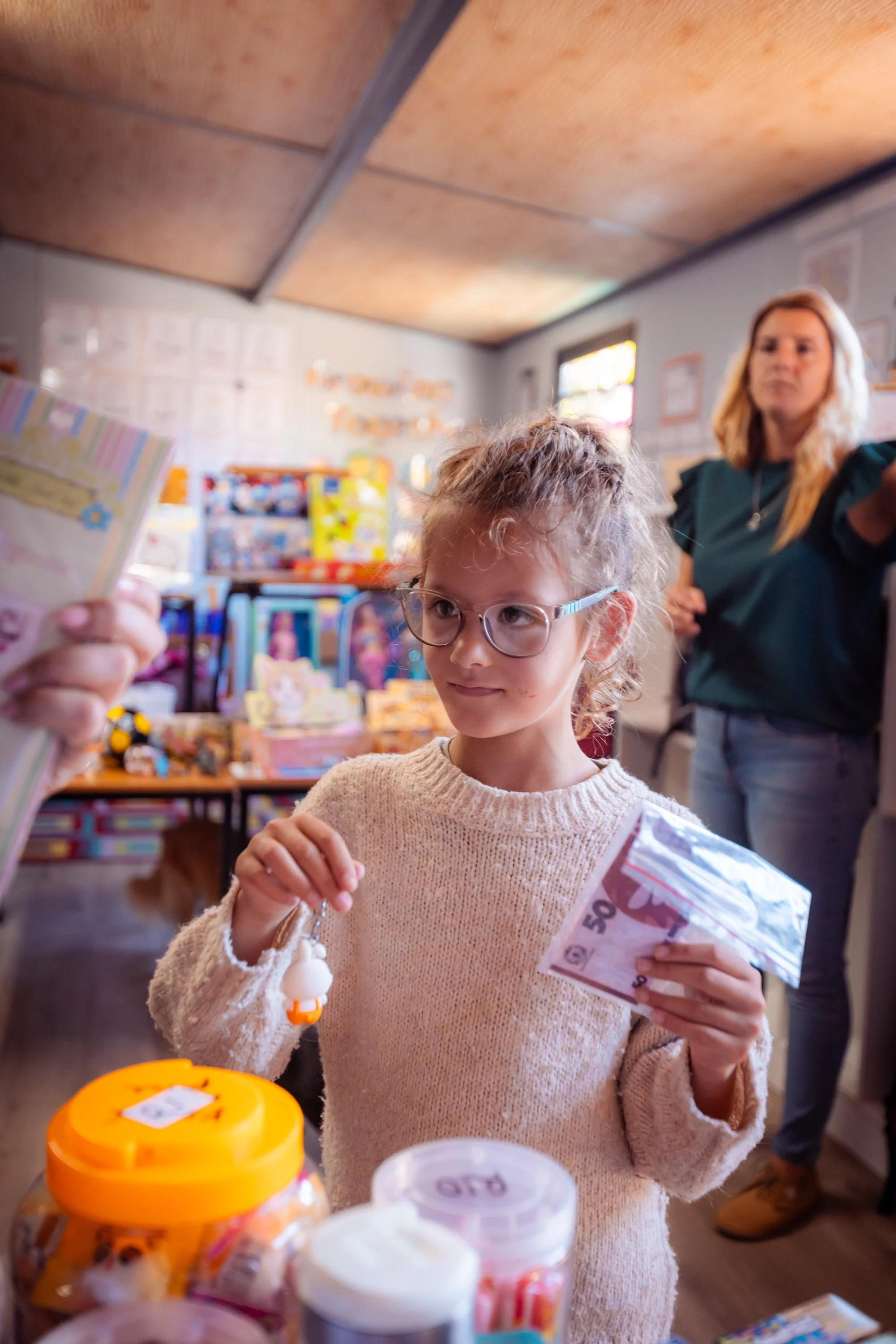 A young girl with glasses and a beige sweater holding a 50 euro banknote while shopping in a store with a woman in the background.