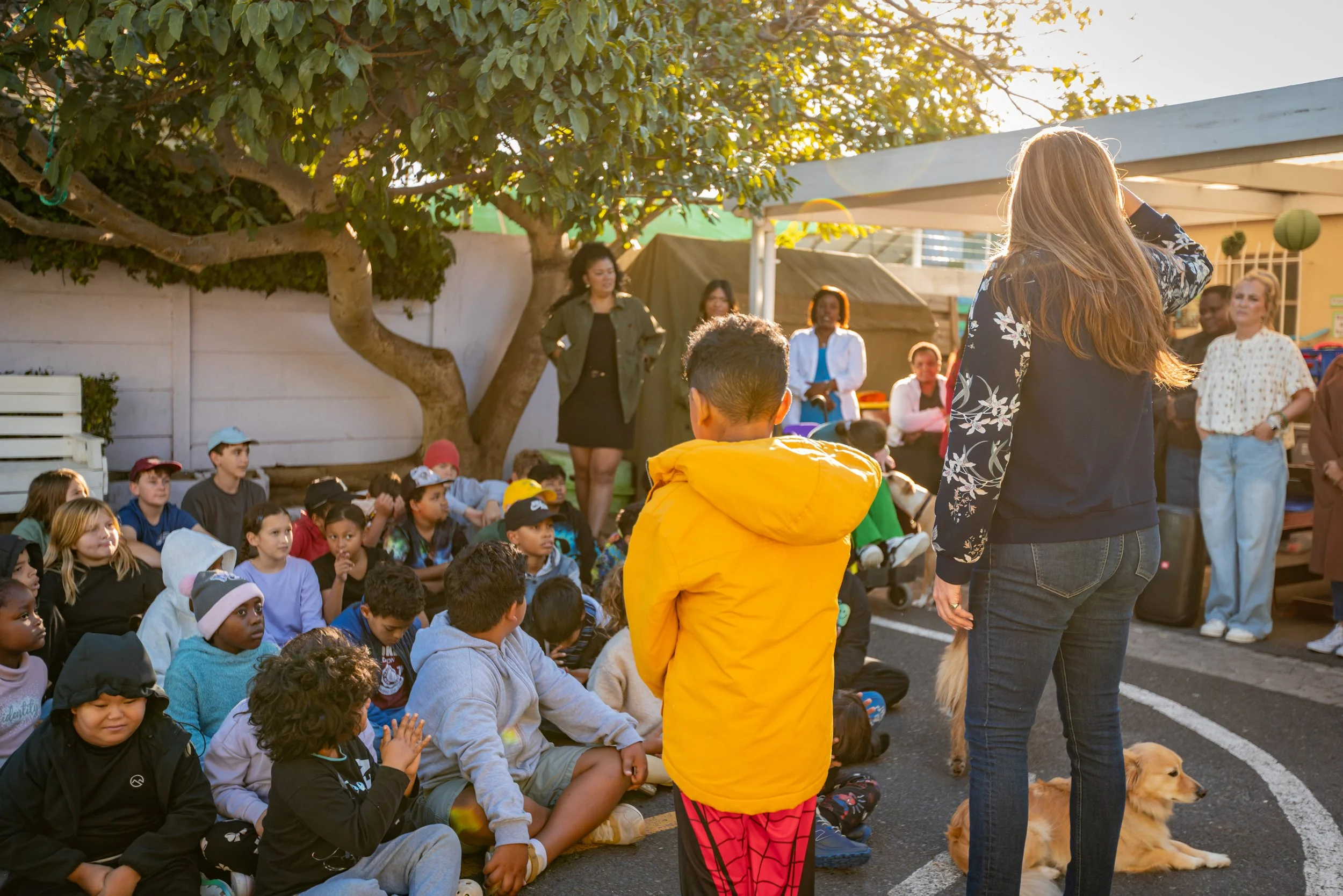 A group of children sitting on the ground, attentively listening to a woman standing in front of them. The woman appears to be speaking or presenting, with several women standing behind her. The scene takes place outdoors under a large tree, with sun