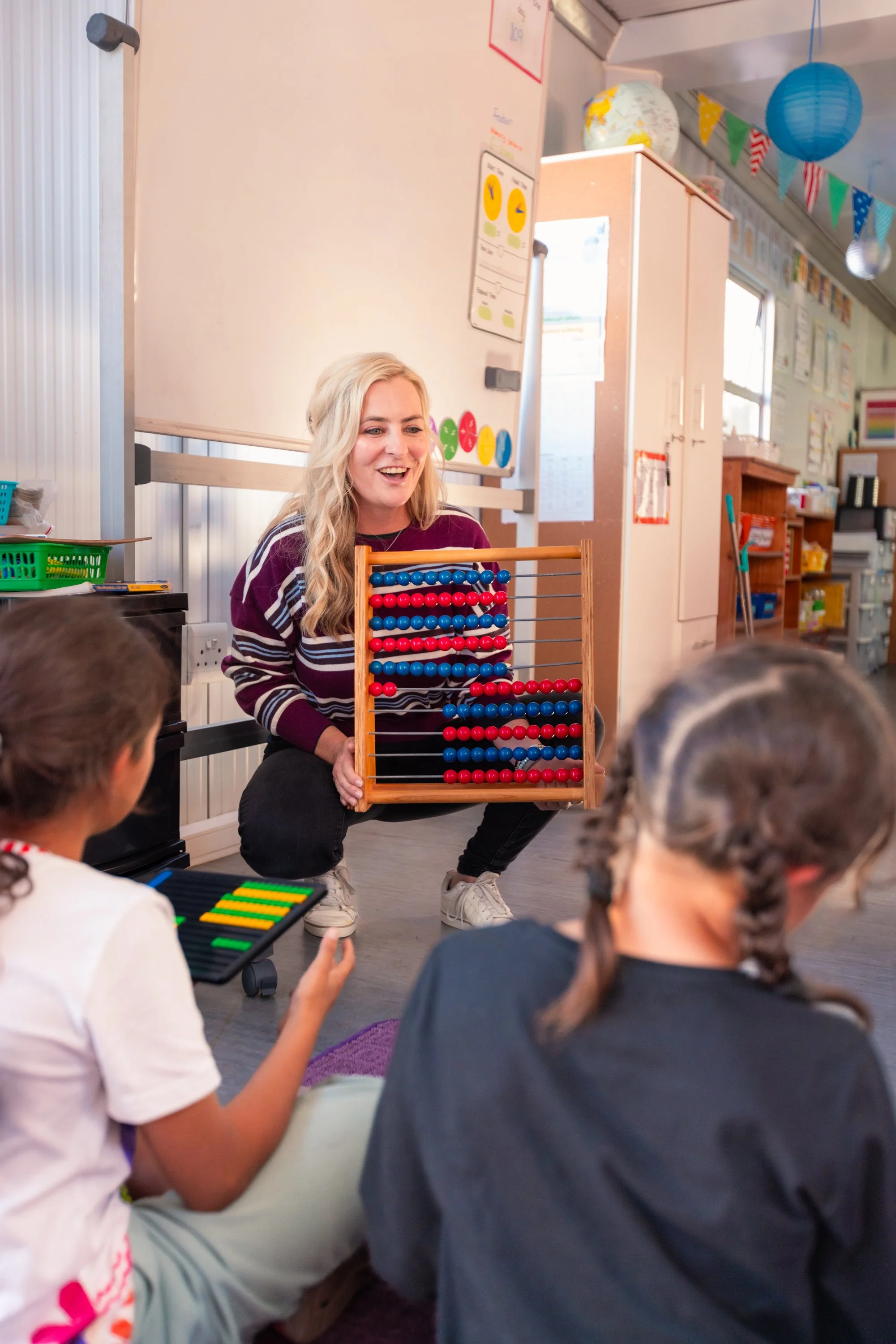 A woman with long blonde hair, wearing a striped maroon and white top, kneeling on the floor, holding an abacus with red and blue beads, engaging with children sitting around her in a classroom decorated with colorful banners and educational material