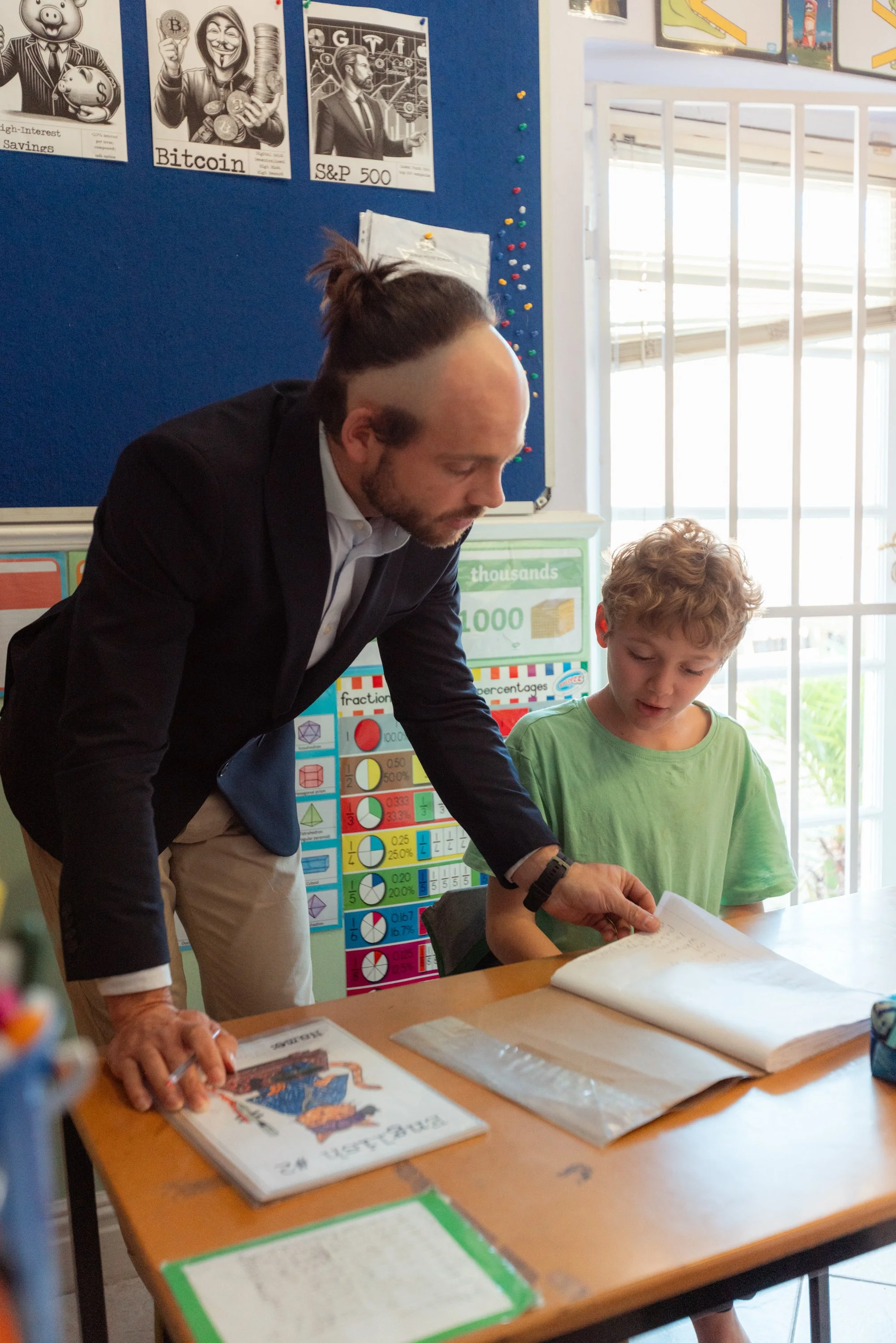A man in a black blazer and beige pants is leaning over a table, pointing at an open book, while a young boy in a green T-shirt looks at the book. They are in a classroom with educational posters about fractions and percentages on the wall.