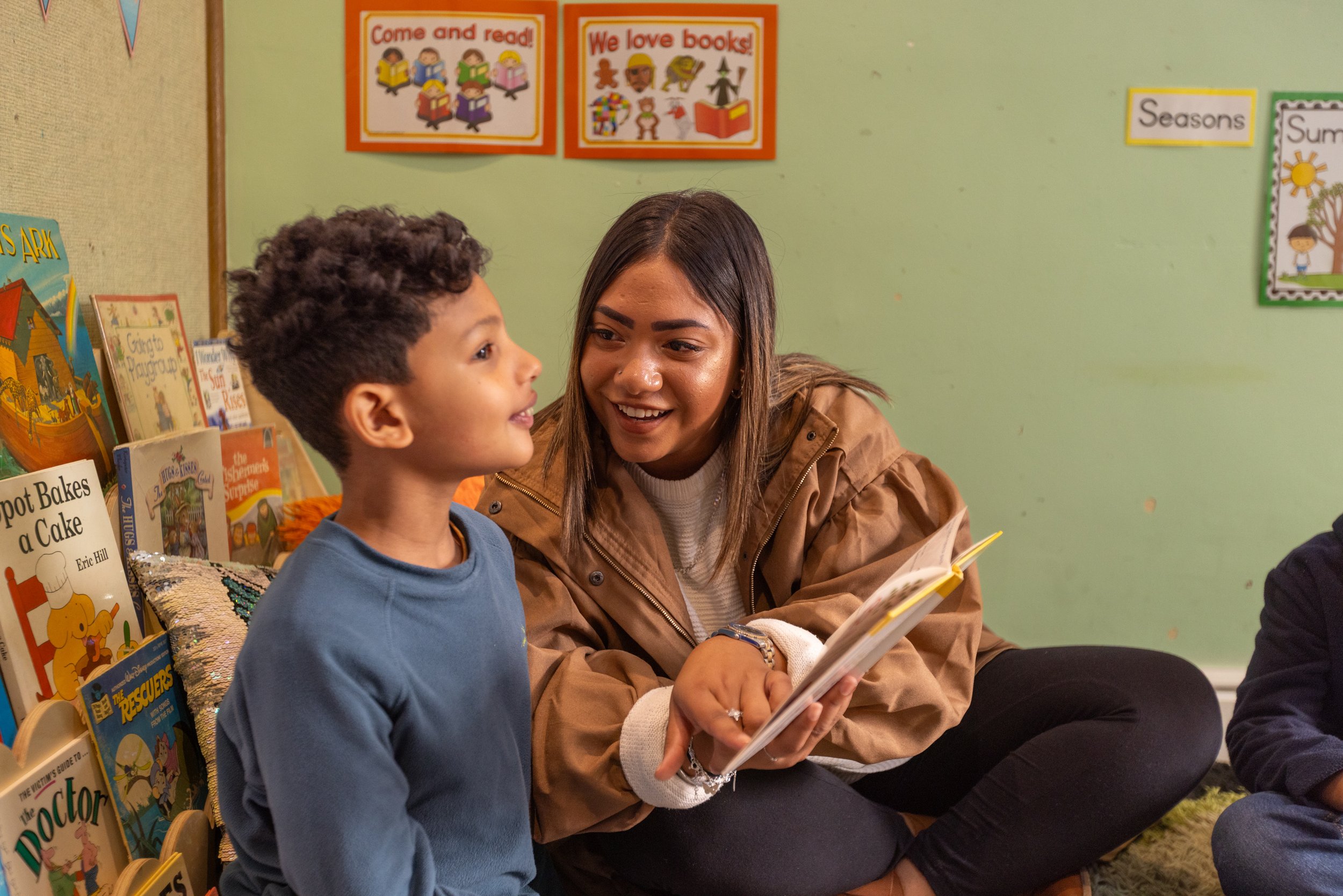 A woman and a young boy are sitting together in a bookstore or library, smiling and looking at a book. Books and colorful posters about reading and seasons are visible in the background.