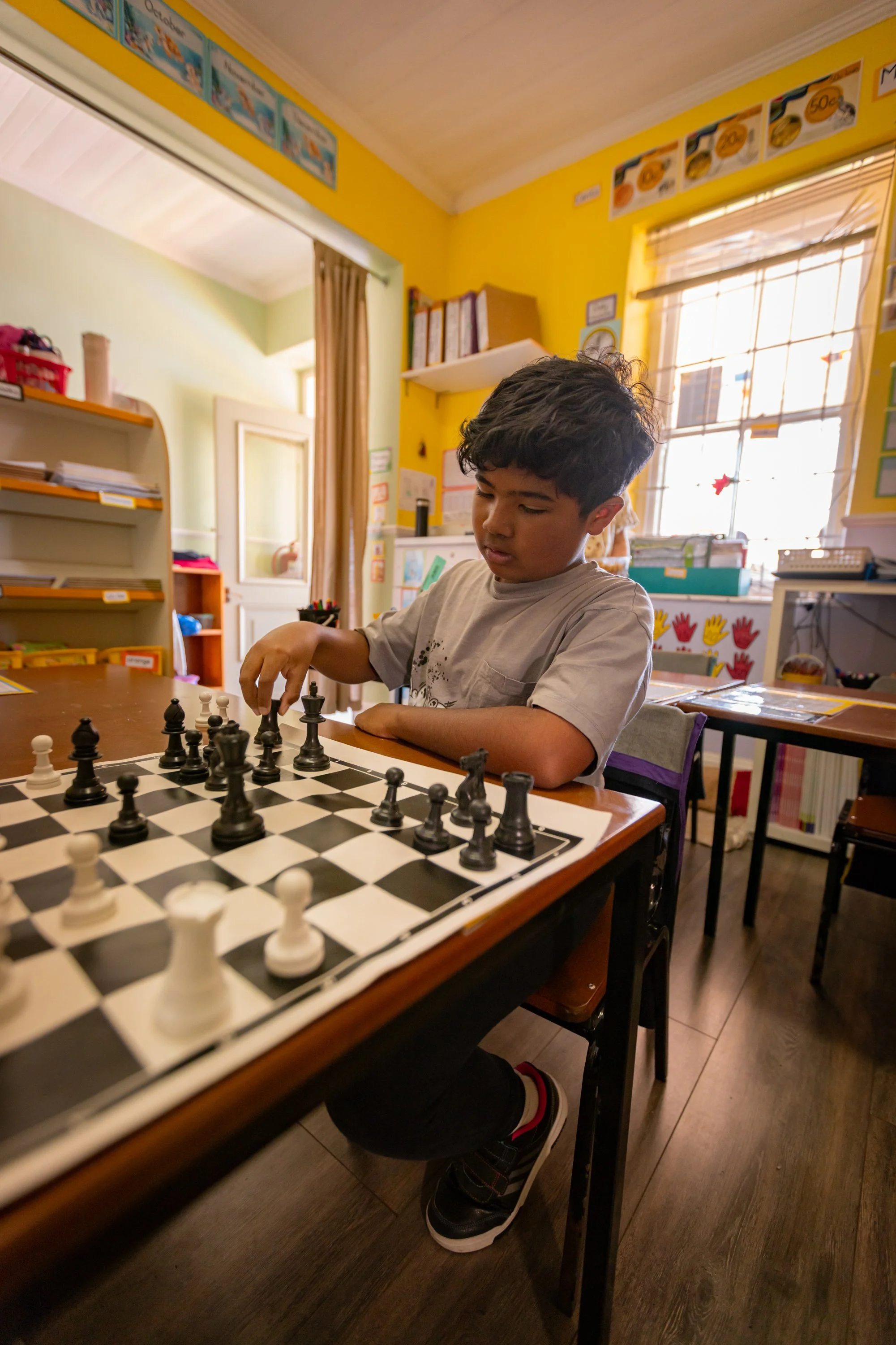 A young boy in a classroom playing chess at a table, with shelves and colorful educational decorations on yellow and green walls, sunlight streaming through a window.