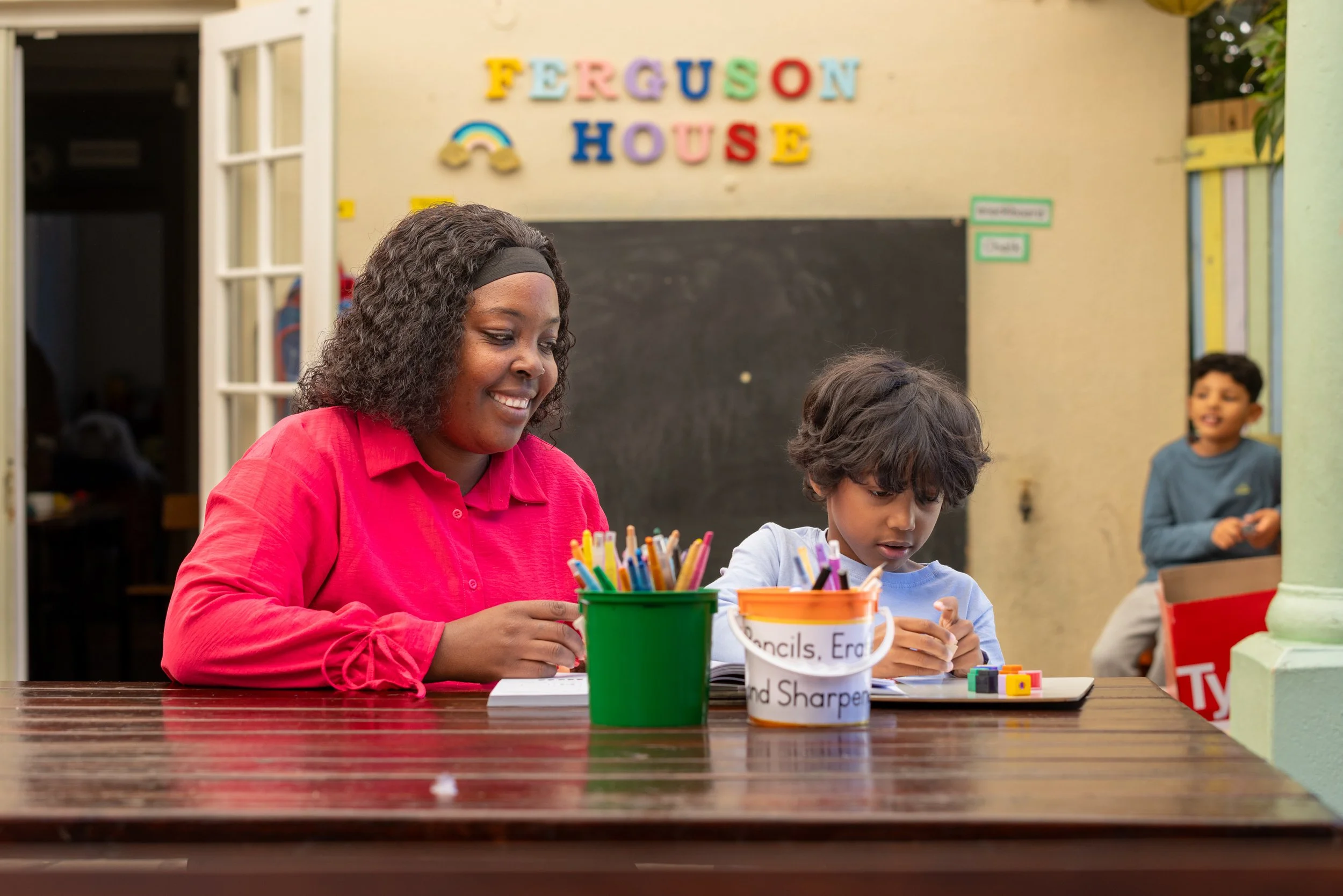 A woman and a young boy sitting at a table, engaging in arts and crafts with colorful markers and paper. In the background, there is a sign that reads 'Ferguson House' decorated with colorful letters and a rainbow. A boy is standing in the background smiling and watching.