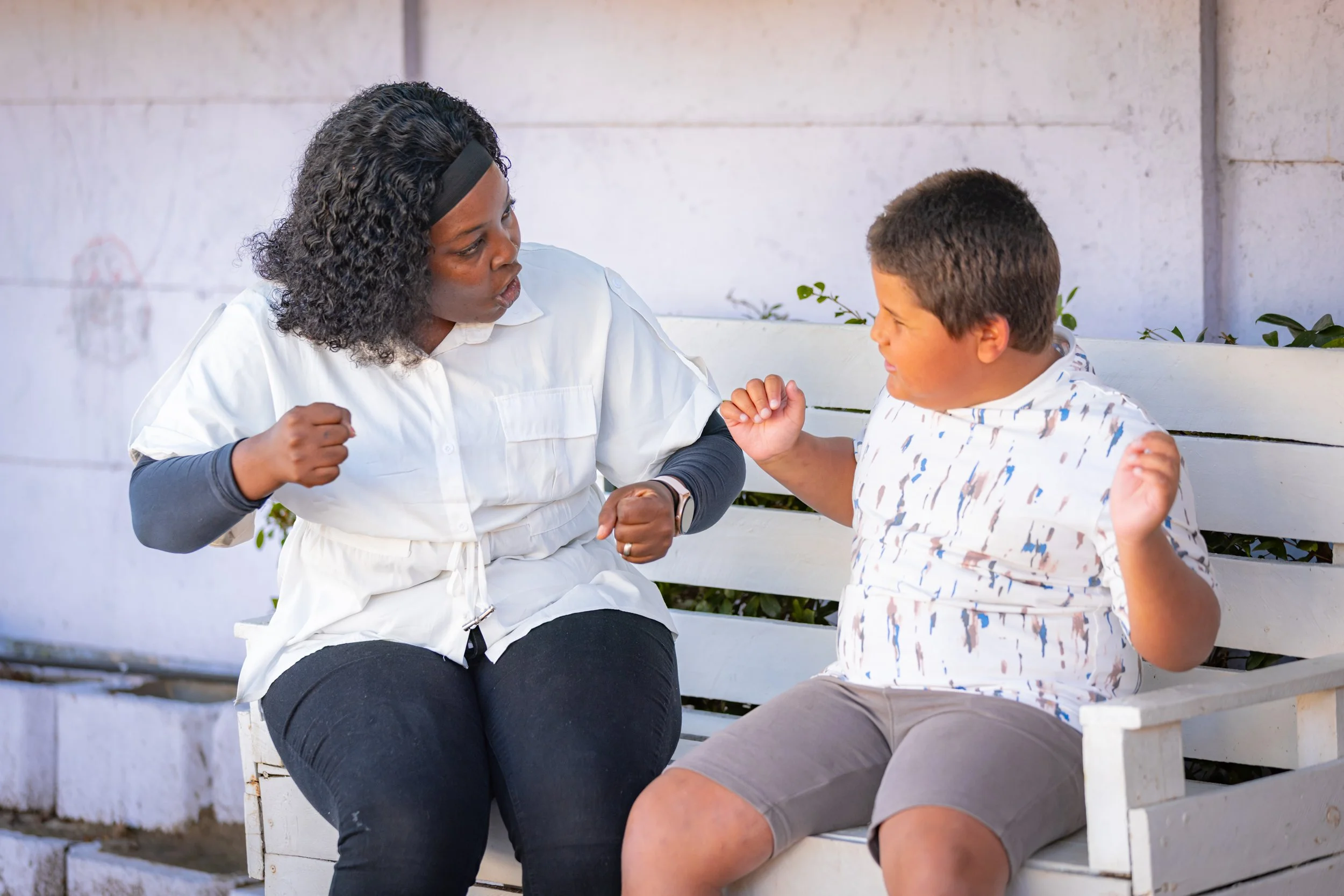 A woman and a boy sitting on a white bench outdoors, having an animated conversation, with the woman making a fist and the boy smiling.