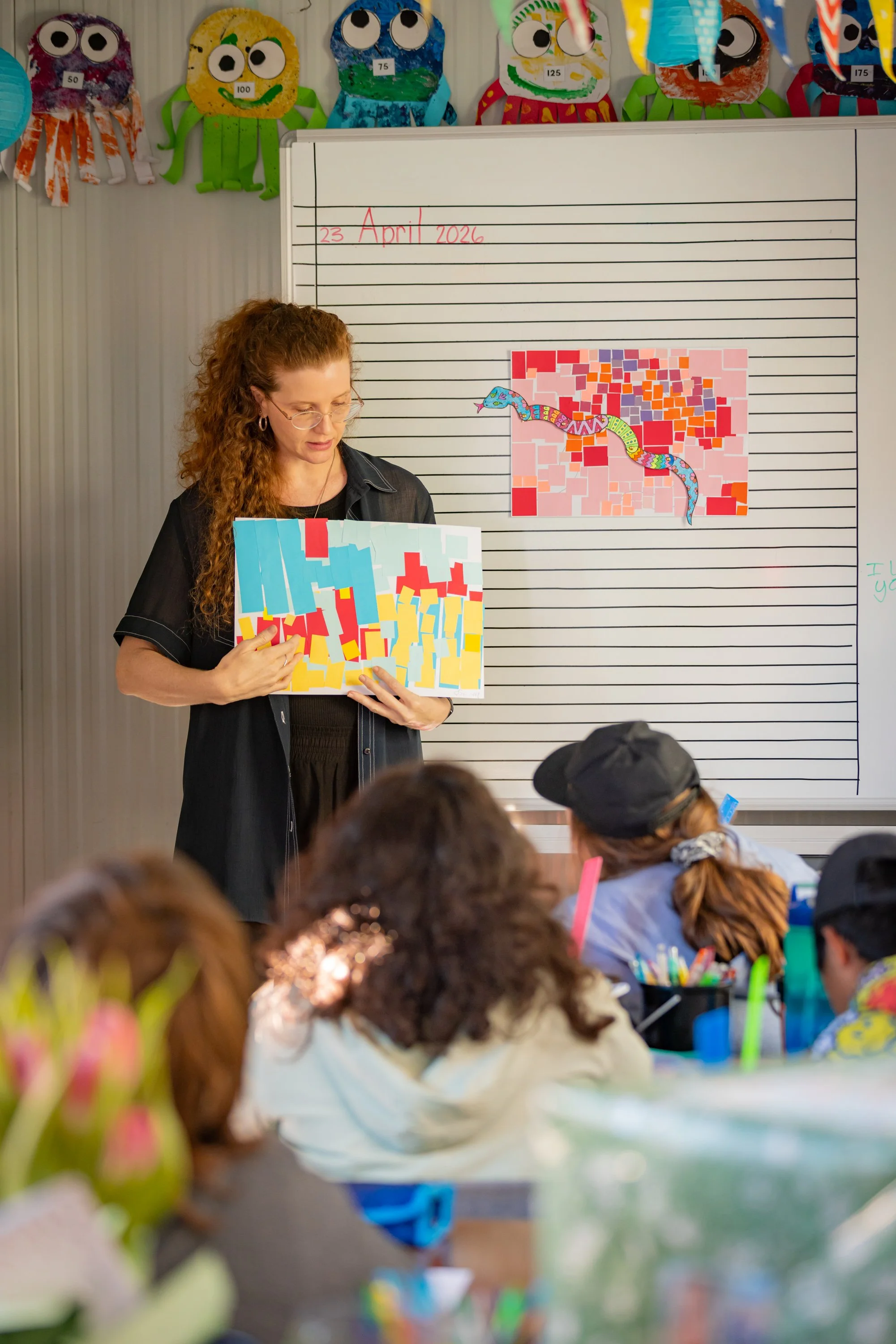 A classroom scene with a woman teacher standing in front of a whiteboard with colorful artwork and writing on it. The whiteboard displays the date '23 April 2026'. Students are sitting at desks, facing the teacher, with some visible in the foreground. The classroom features paper octopus decorations on the wall above the whiteboard, each with a different number. The students are engaged, some with notebooks and pens.