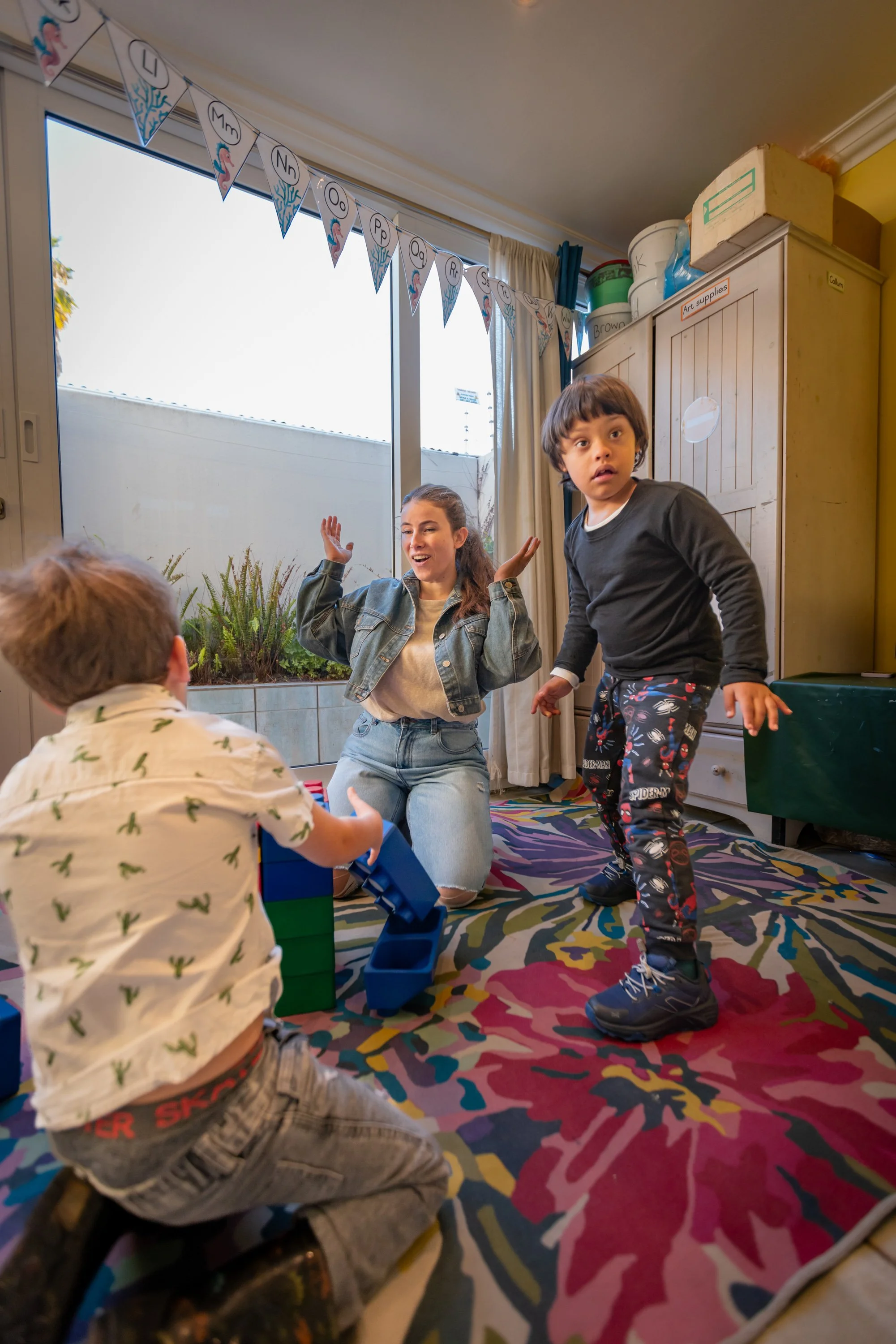 Children and a young woman in a colorful room with a rainbow carpet, engaging in a playful activity near a window decorated with a banner of the alphabet. A cabinet labeled 'Art supplies' is in the background.