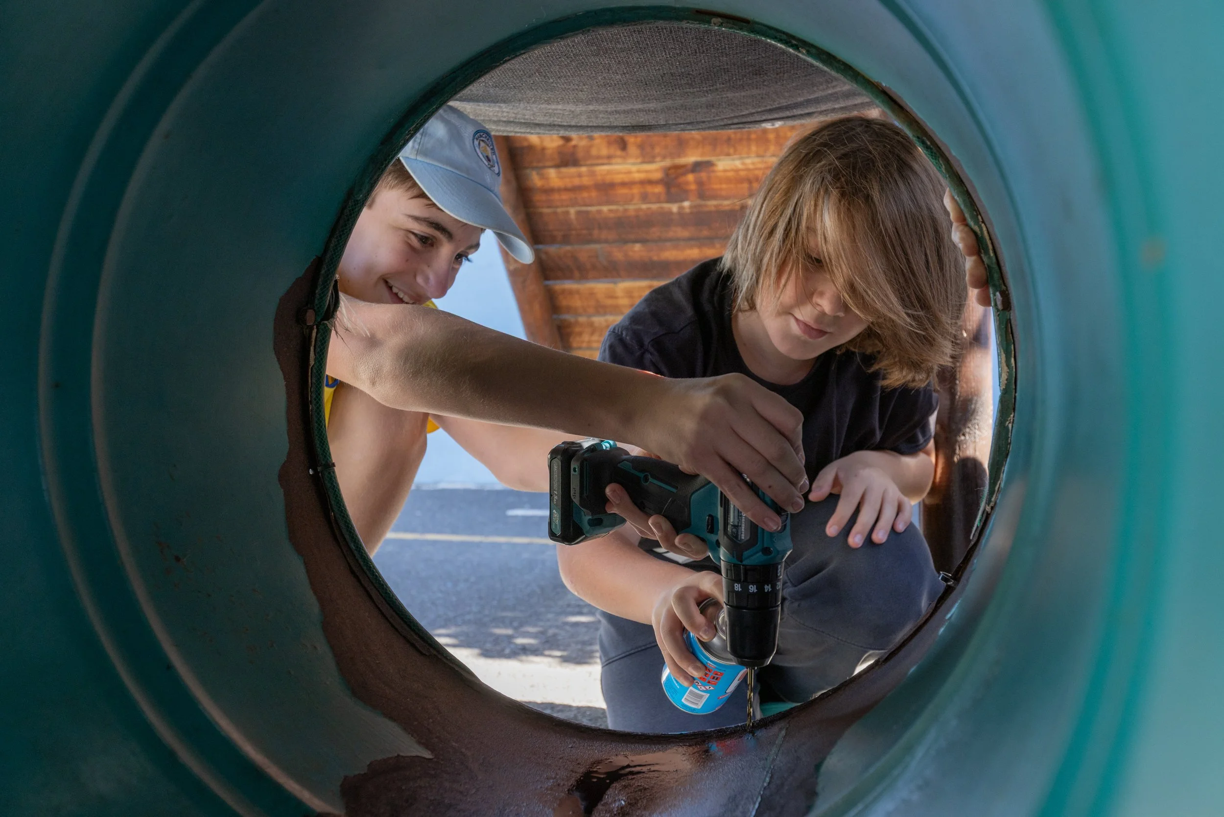 Two boys with long hair and one in a blue cap are inside a green pipe, using a drill to fix or adjust something.