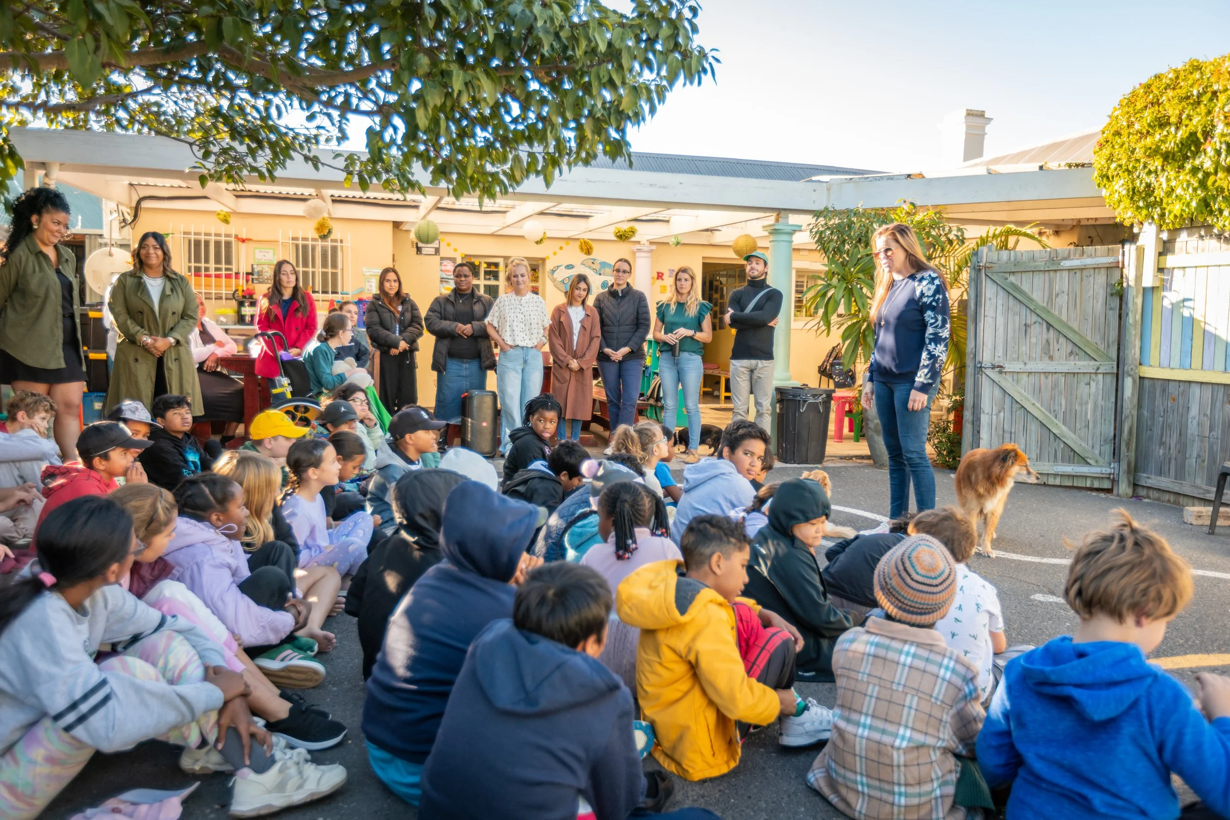 A group of children seated on the ground outdoors listening to a woman speaking. Several adults are standing behind them in a backyard setting decorated with paper lanterns and streamers, with trees and a building in the background.