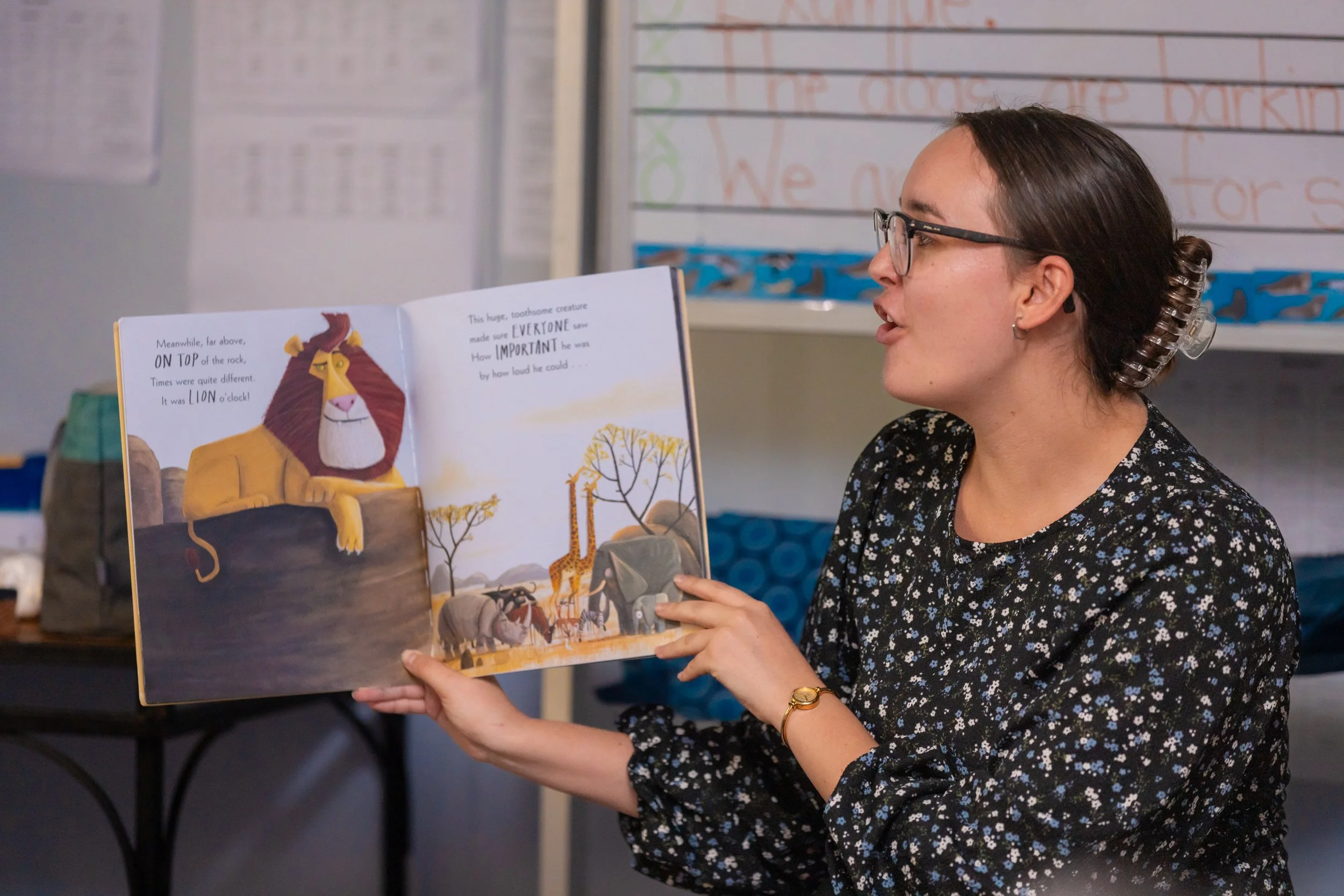 A woman with glasses, earrings, and a black floral dress is reading a children's picture book with a lion and other animals in an illustrated scene. She has a hair clip and a gold bracelet. The background shows a classroom setting with a whiteboard a