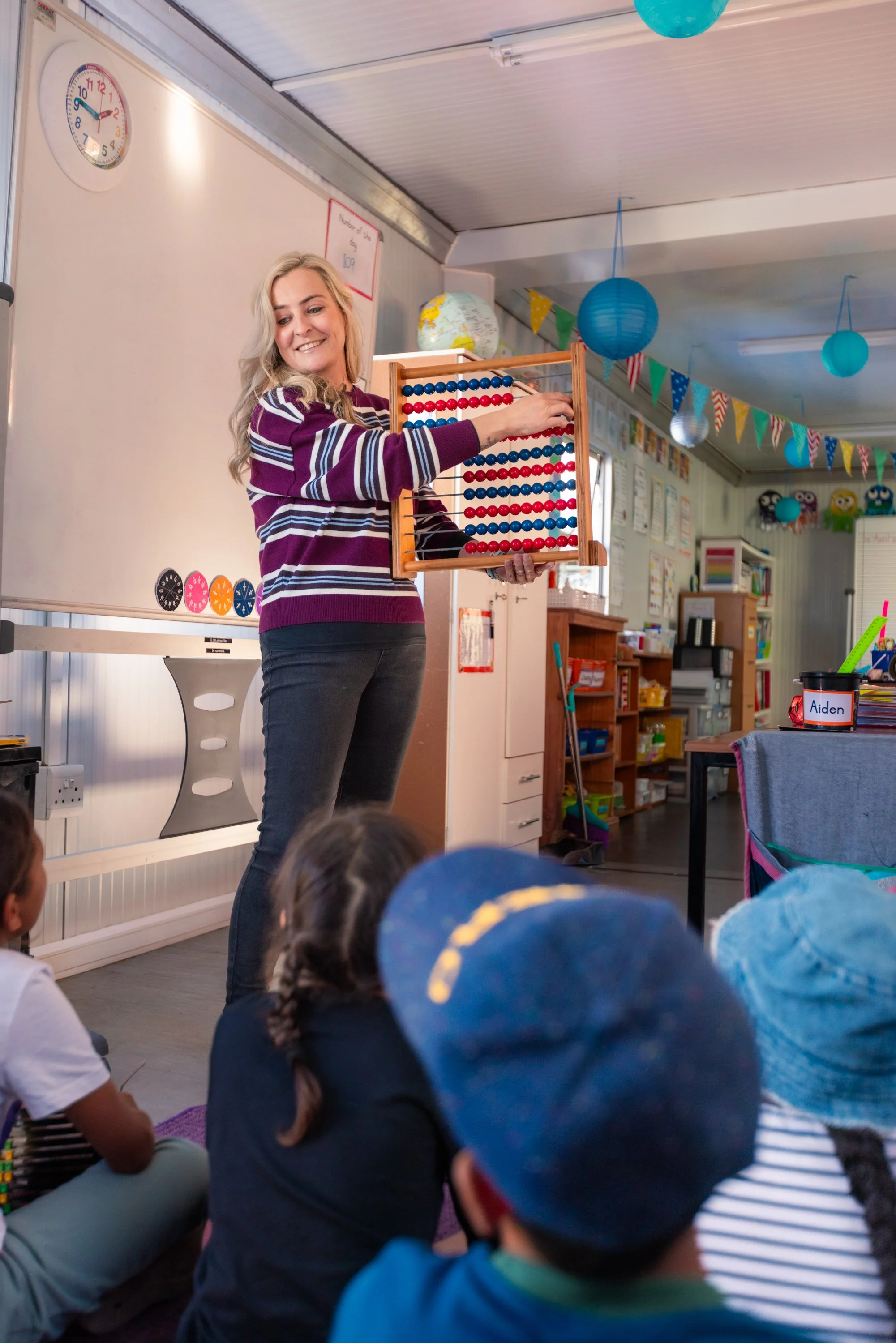 A woman with blonde hair in a striped purple and white sweater is standing and smiling while holding a large wooden abacus with red and blue beads in a classroom decorated with colorful hanging paper lanterns, paper bunting, and educational posters.