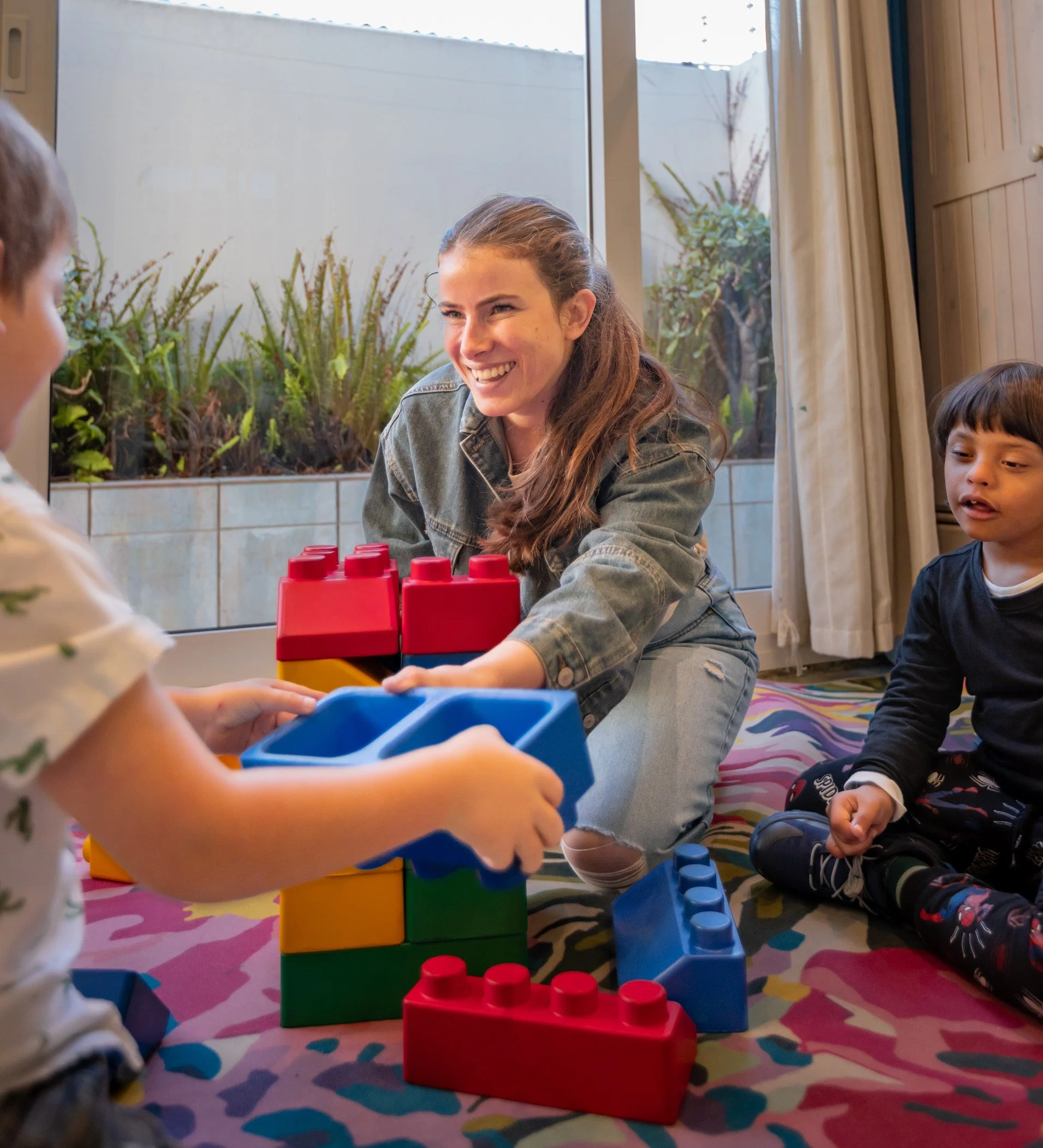 Children playing with large colorful toy building blocks inside house near window, with plants outside