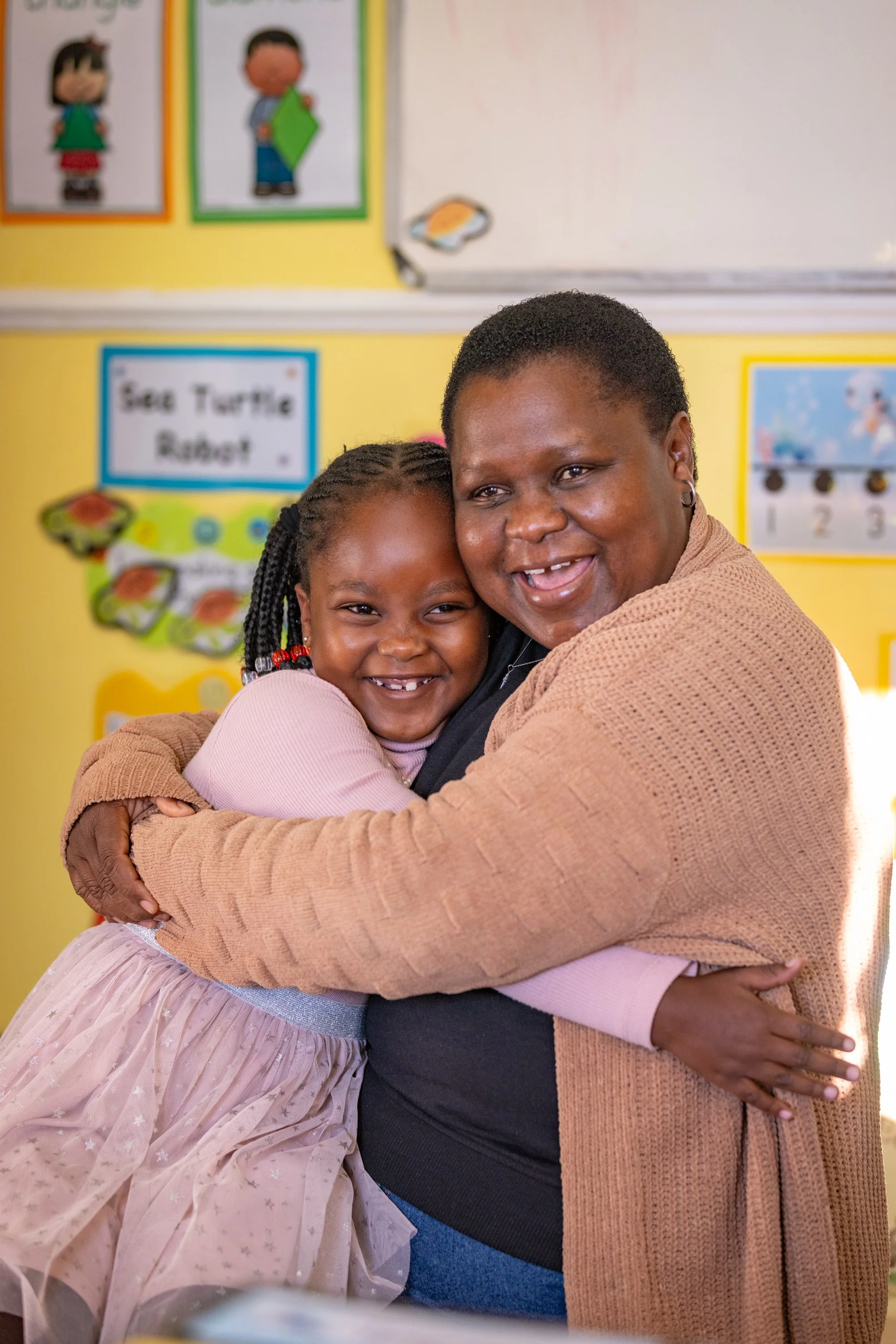 A woman and a girl hugging and smiling in a classroom with educational posters on the wall.