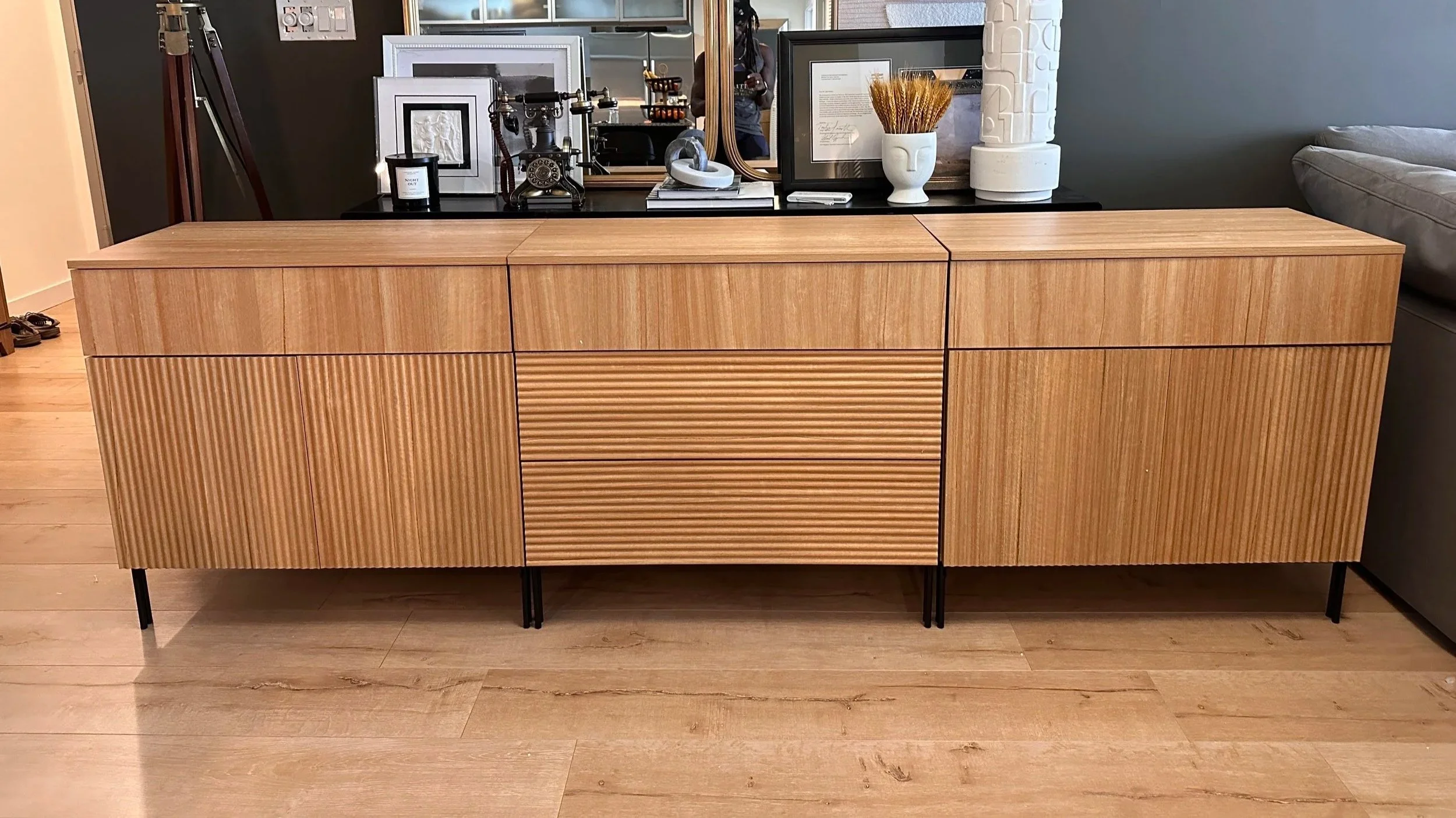 A modern wooden sideboard with a matte black top, featuring vertical and horizontal grooved wood panels, and supported by black metal legs, situated on a light wooden floor.