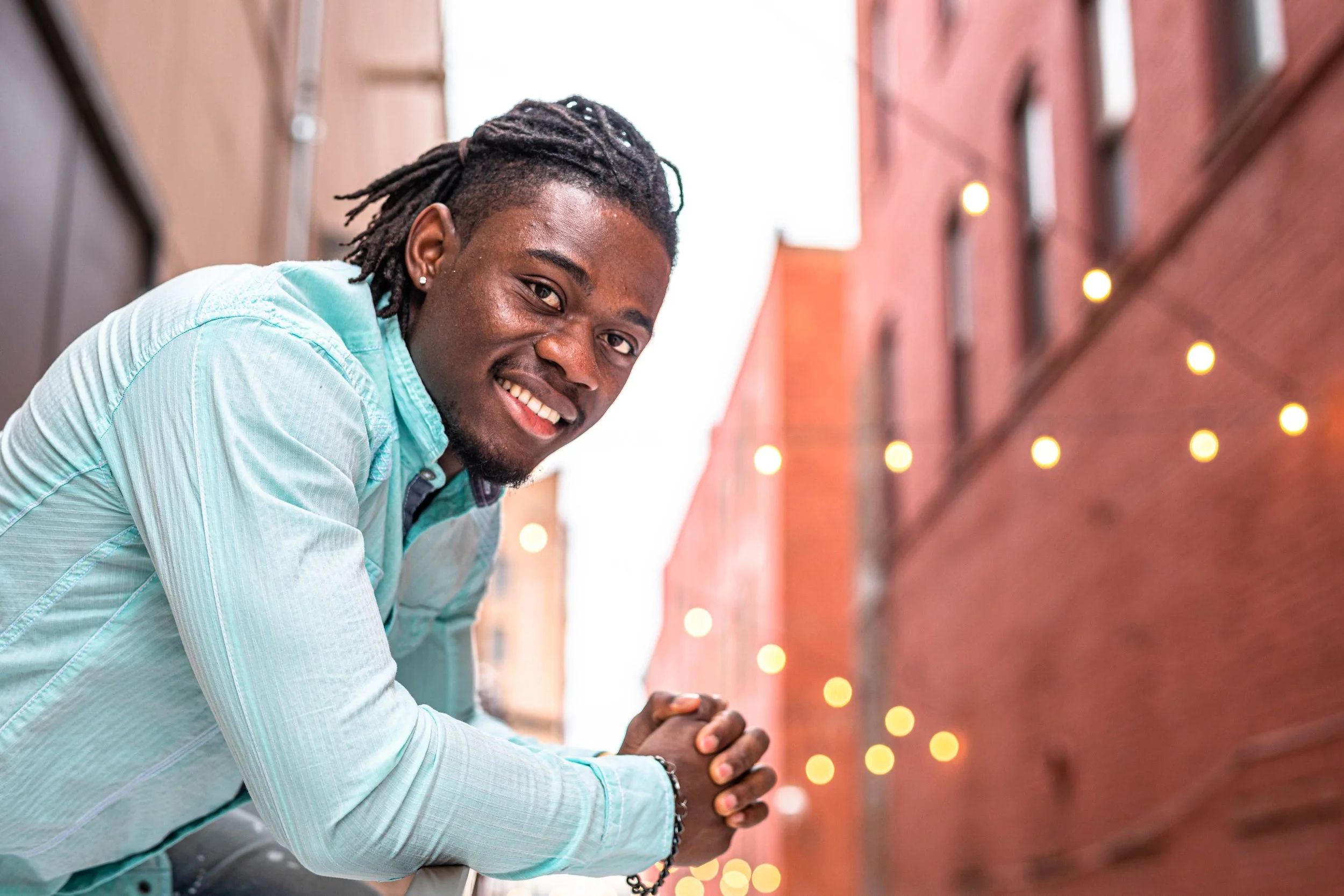 A young man with dreadlocks smiling and leaning on a brick wall with string lights in the background, wearing a light blue shirt.