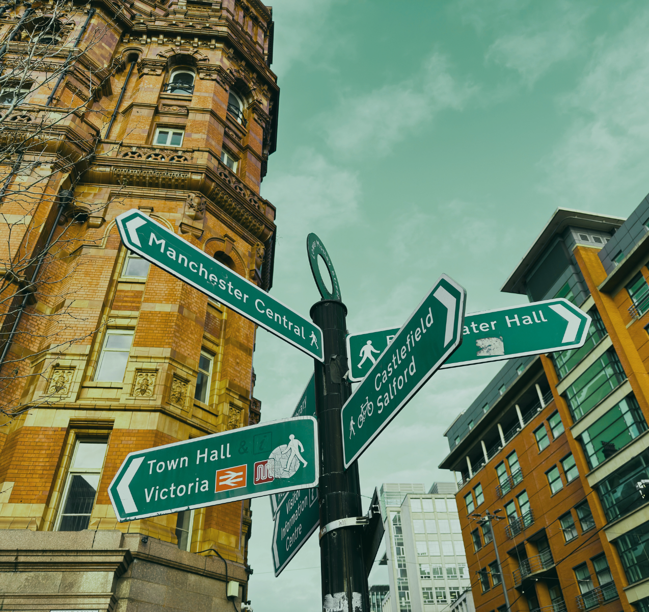 Street signs at an intersection in Manchester, UK, showing directions to Manchester Central, Castlefield, Salford, Town Hall, Victoria, and Water Hall, with historic and modern buildings in the background under a cloudy sky.