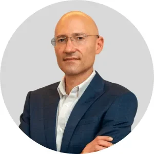 Uomo con abito blu e camicia bianca, con braccia incrociate, sfondo grigio chiaro.