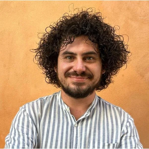 Ragazzo con capelli ricci, barba e maglietta a righe in piedi davanti a una parete arancione.