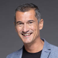 Primo piano di un uomo sorridente con capelli corti e grigi, che indossa una giacca blu e una maglietta nera, sfondo grigio