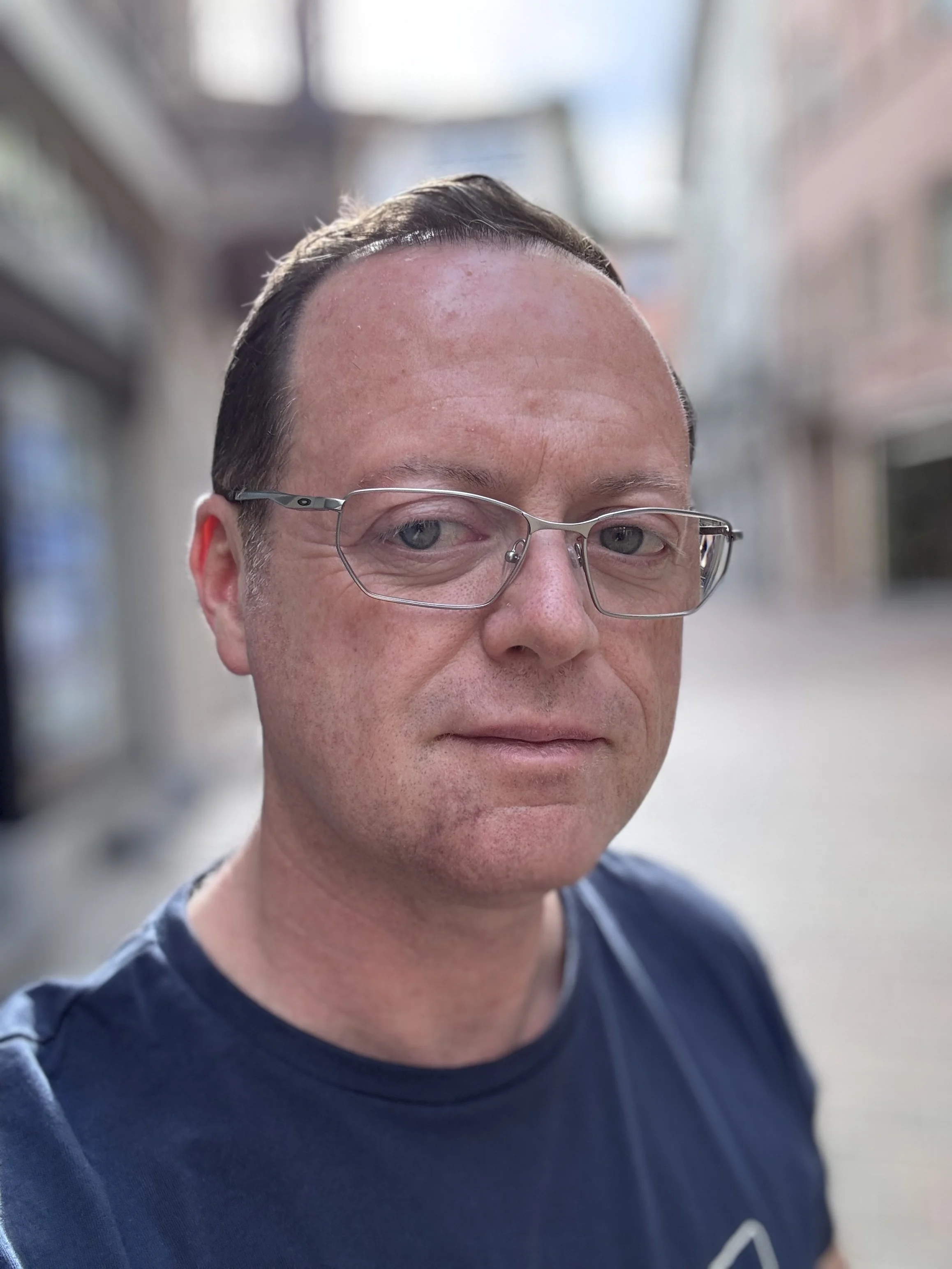 Ritratto di un uomo con occhiali, capelli corti e scuri, in vestiti casual, su strada con edifici sullo sfondo.