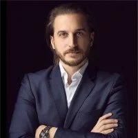 Ritratto di un uomo con capelli castani e barba, indossa un completo blu e camicia bianca, sfondo scuro.