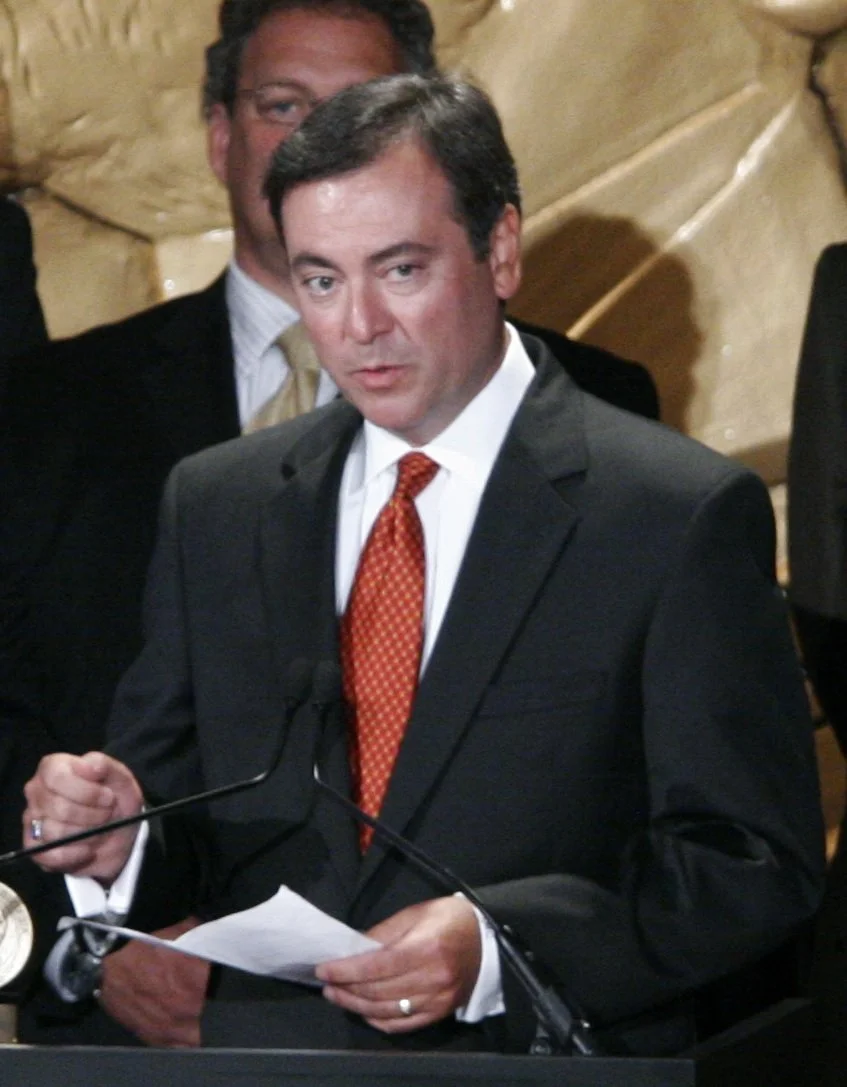Man in a black suit wearing a red tie speaking at a podium during a formal event, with another man in a suit standing behind him.