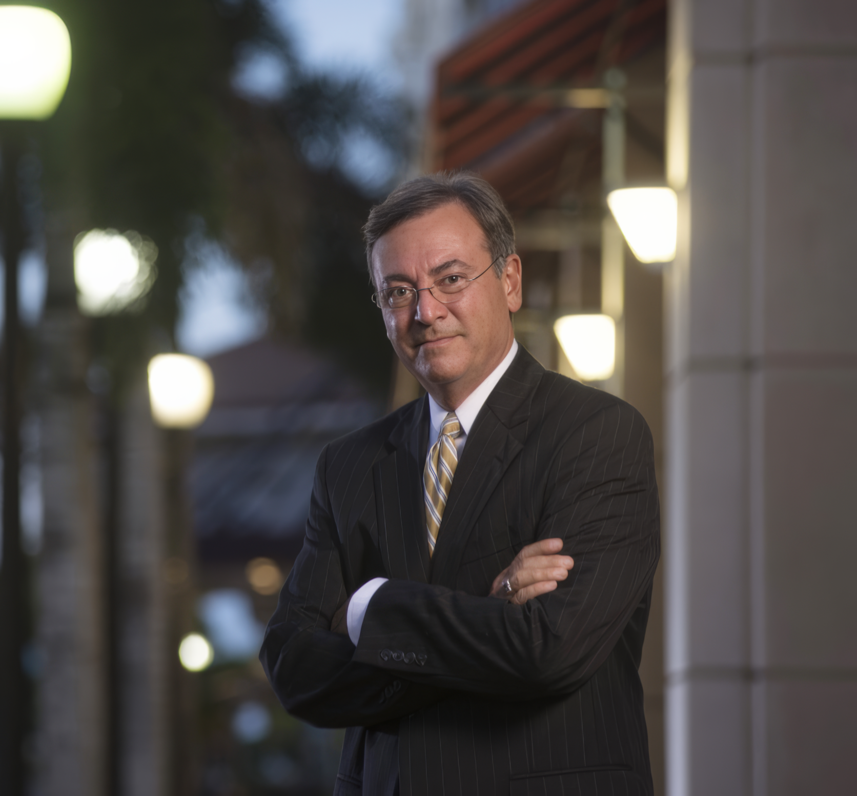A middle-aged man in a black pinstripe suit with a light-colored striped tie, glasses, and short gray hair, standing outdoors at dusk, with arms crossed and a confident expression, in front of a building with exterior lights.