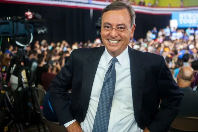 A man in a suit smiling at an indoor event with a large audience in the background.