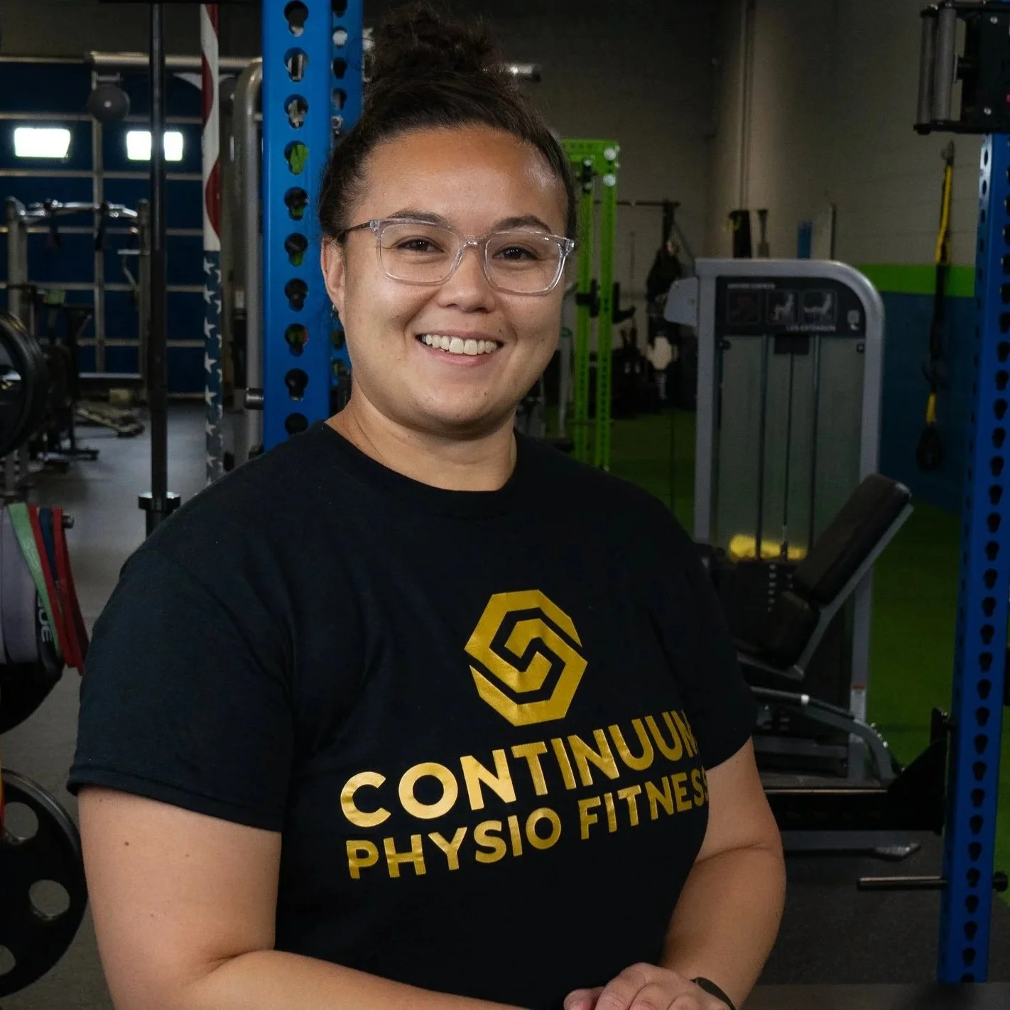 A woman with glasses and her hair in a bun smiling at the camera inside a gym. She is wearing a black t-shirt with yellow text that reads "CONTINUUM PHYSIO FITNESS" and a yellow logo on her shirt. The background features gym equipment including weight racks, a workout machine, and green and blue gym bars.