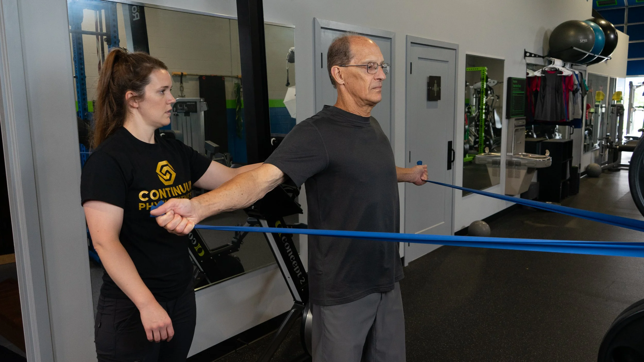 A young woman assisting an elderly man with resistance band exercises in a gym, with workout equipment visible in the background.